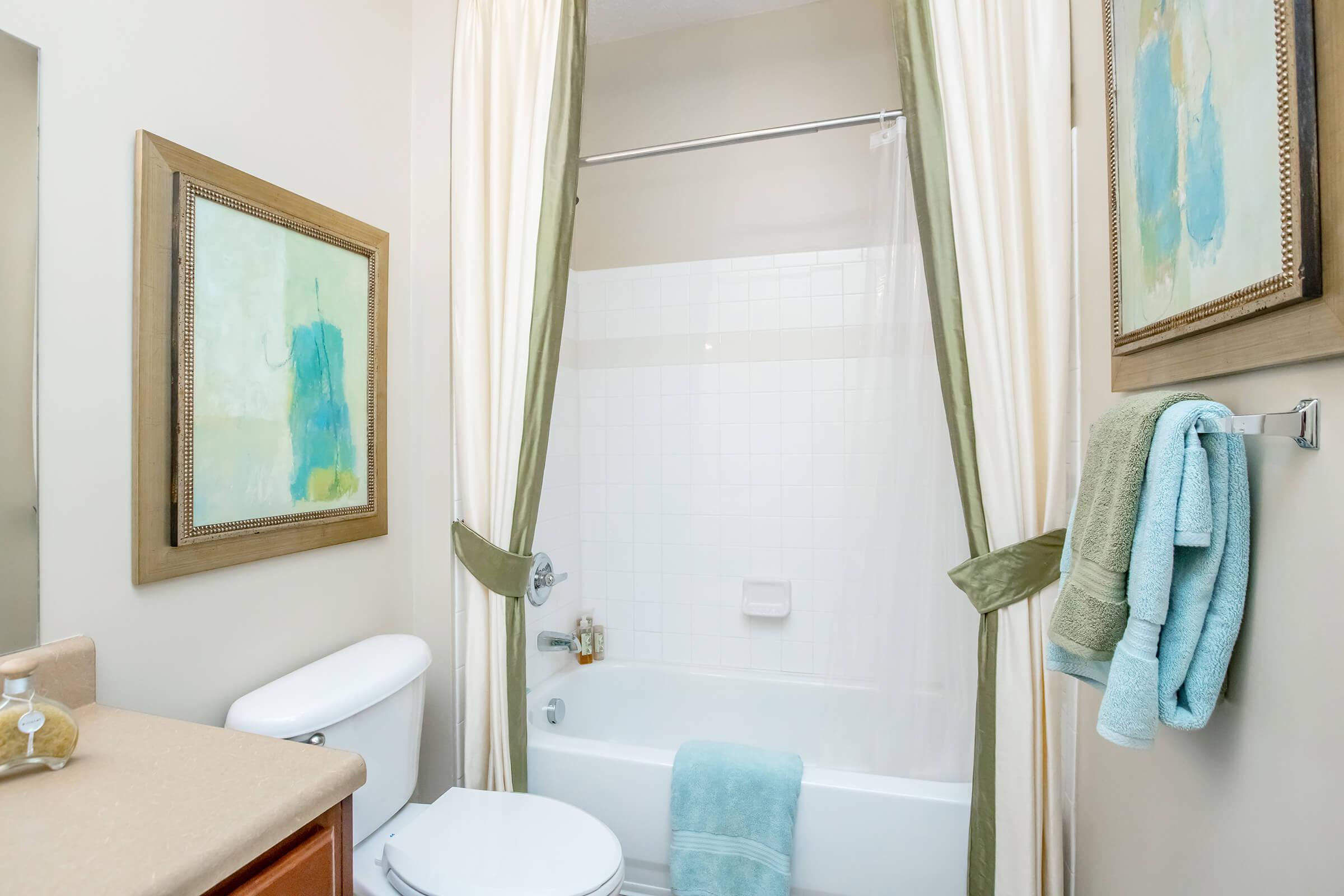 A clean bathroom featuring a bathtub with a shower curtain, light green accents, a framed abstract painting on the wall, and neatly arranged towels. The vanity has a beige countertop and a sink, contributing to a bright and inviting atmosphere.