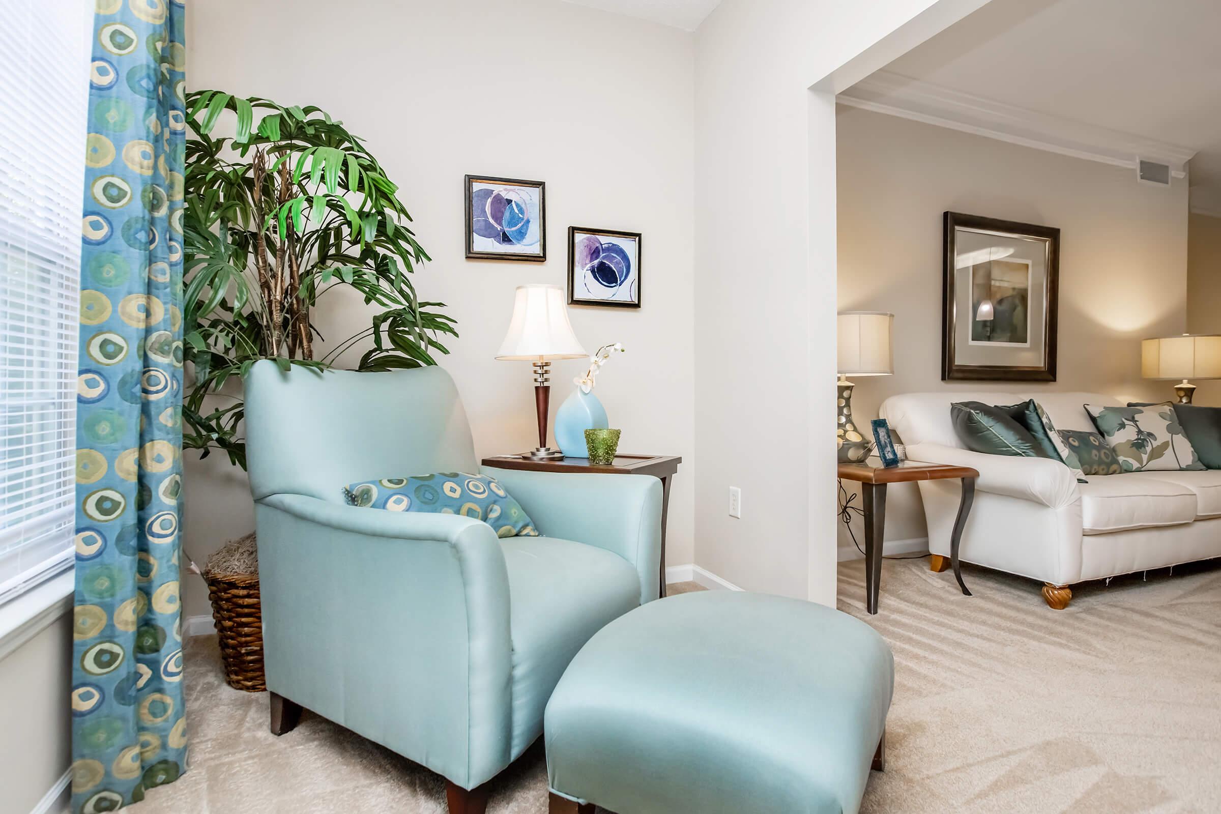 A cozy living room corner featuring a light blue armchair and matching ottoman, adorned with decorative cushions. There are two lamps on either side, and a small table with a vase. In the background, a white sofa with pillows and framed artwork on the wall, complemented by a potted green plant.