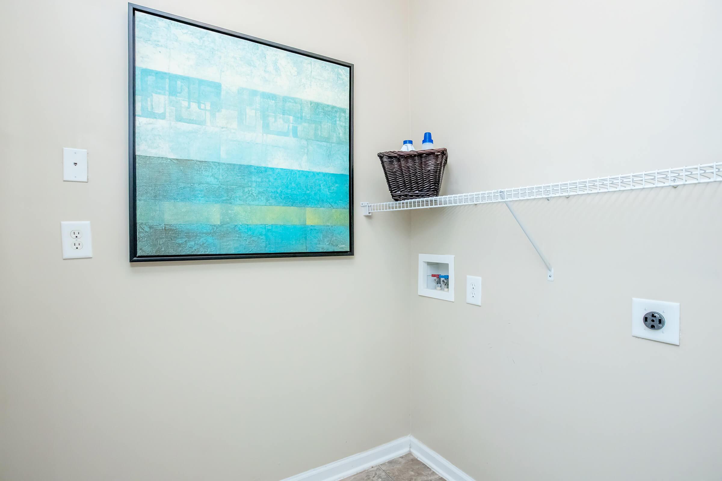 A simple laundry room featuring a light-colored wall, a large framed abstract painting in shades of blue, and a wire shelf holding a wicker basket. There are electrical outlets on the walls, and a small framed item is mounted beneath the shelf. The flooring is a neutral tone.