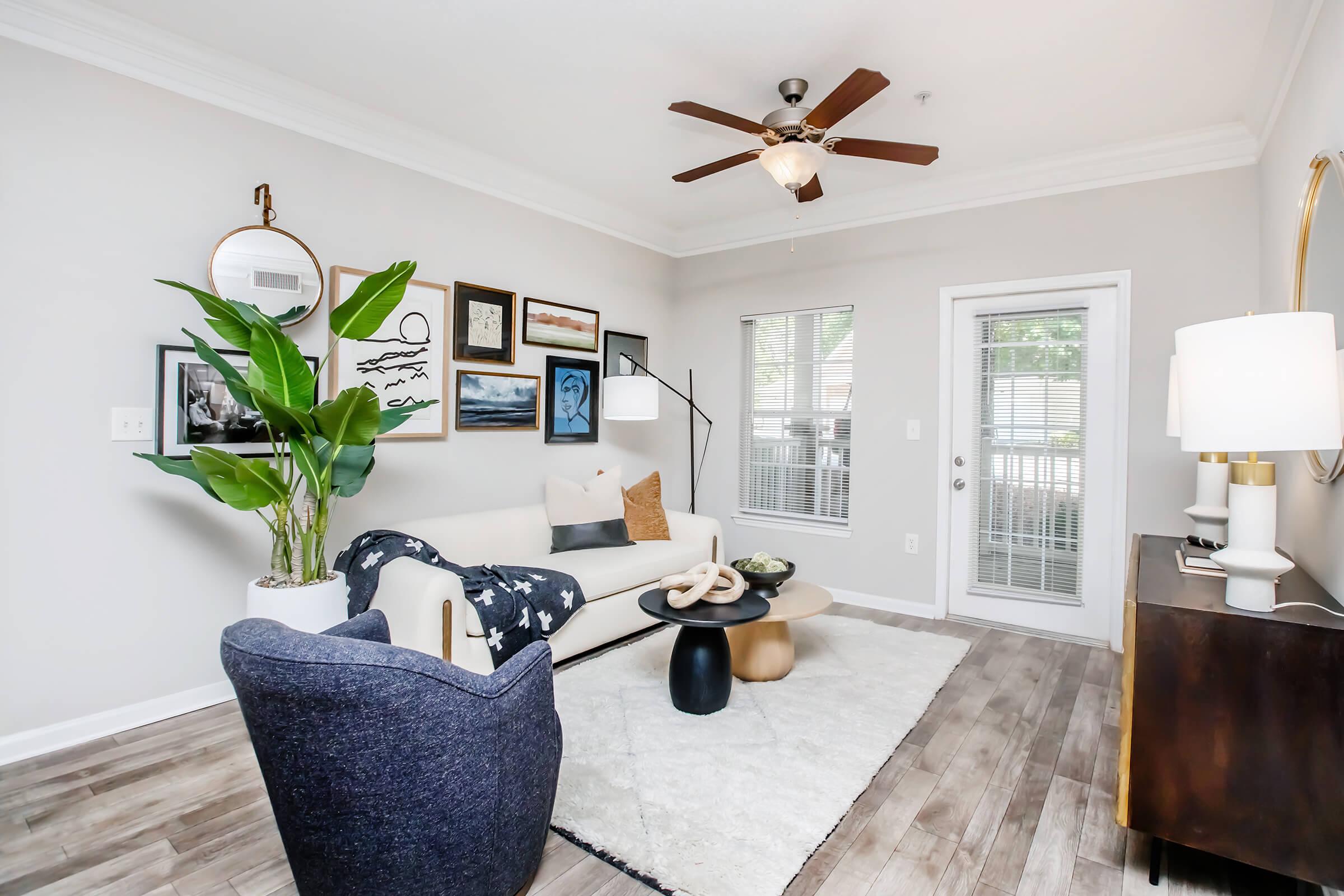 A stylish living room with light gray walls, featuring a white sofa with decorative pillows, a dark blue armchair, and a round white rug. Artwork adorns the walls, and a tall plant adds greenery. A ceiling fan provides airflow, with large windows allowing natural light. A modern lamp and console table complete the decor.