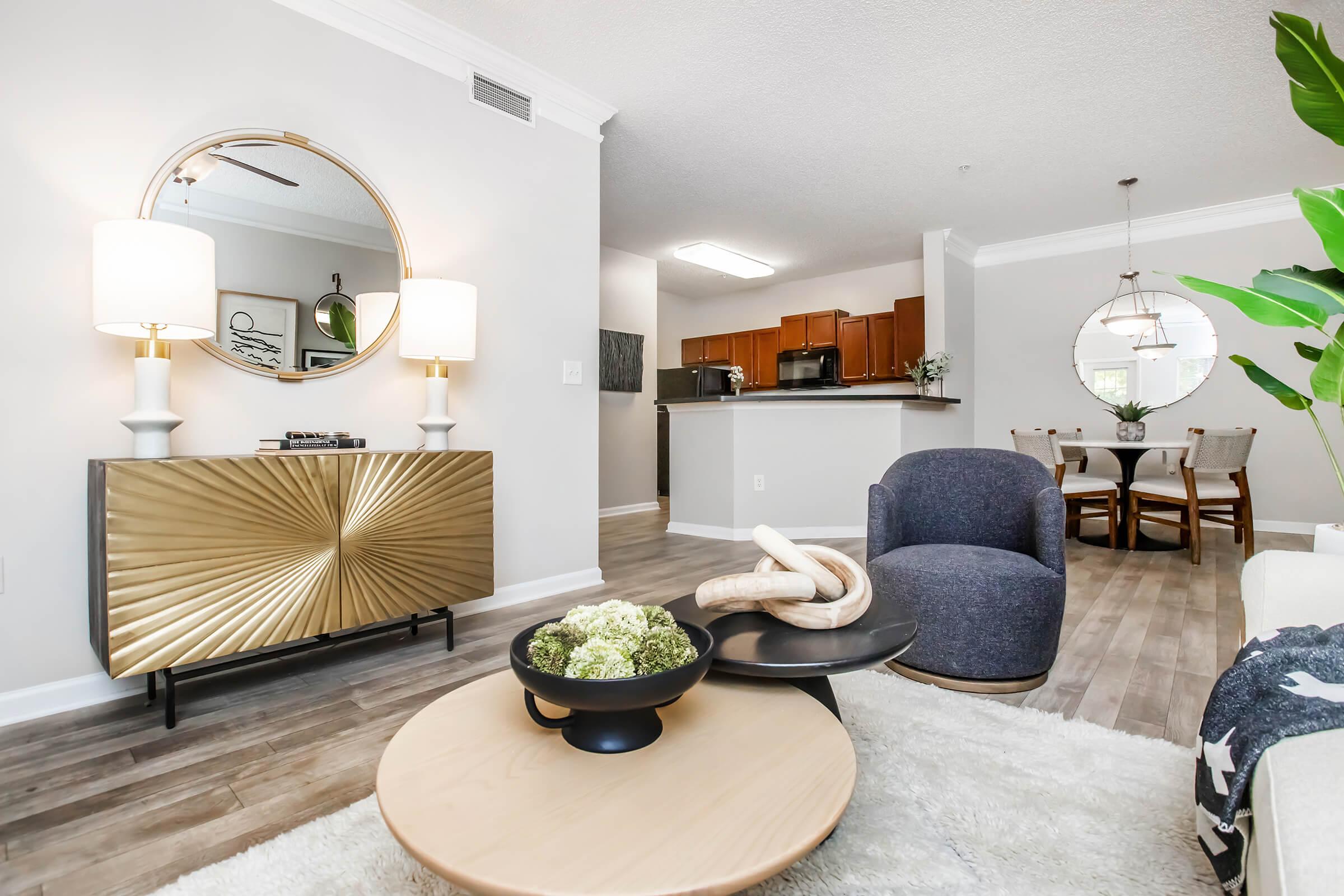 Modern living room with a stylish gold-accented console table, two lamps, and a round mirror. A cozy blue armchair and a circular coffee table with decorative items are in the foreground. In the background, an open kitchen with wooden cabinets and a dining area featuring a circular table can be seen.