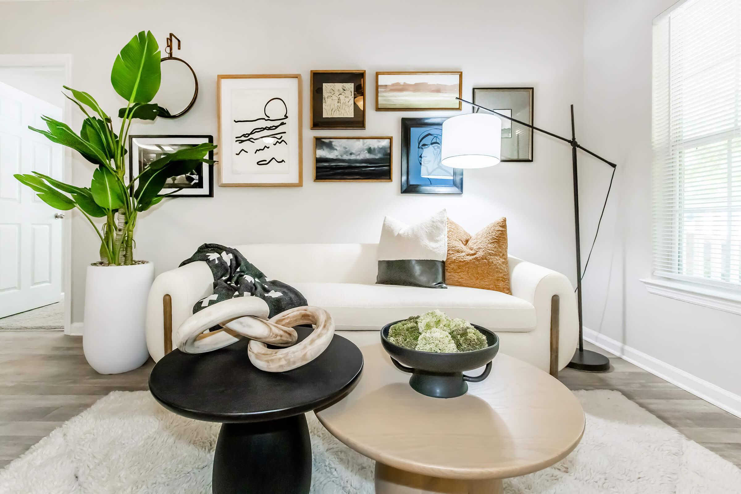 A cozy living room featuring a white sofa adorned with decorative pillows, a round coffee table with a black bowl filled with greenery, and another circular table. The walls are decorated with various framed artworks. A large green plant adds a touch of nature, while a floor lamp provides soft lighting.