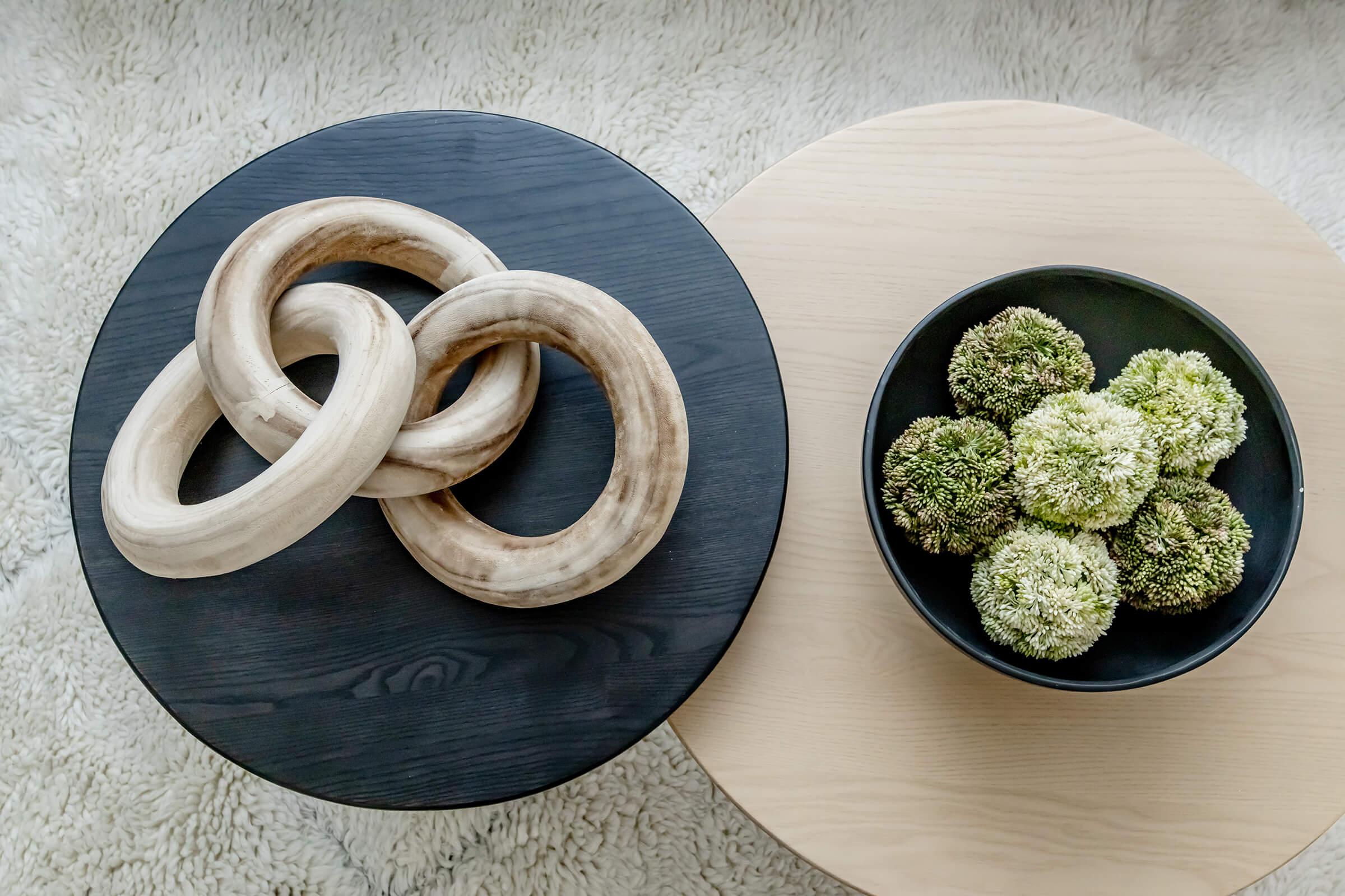 A wooden sculpture consisting of intertwined rings on a black circular table, alongside a bowl filled with green moss spheres on a lighter circular table. The surfaces are complemented by a soft, textured rug underneath.