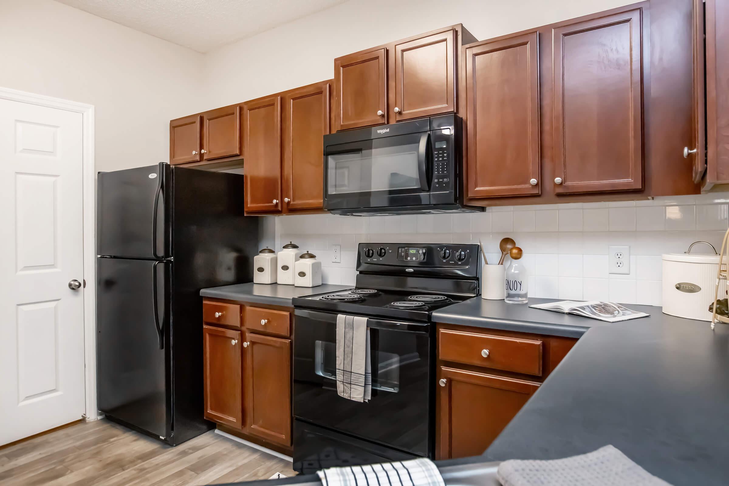 A modern kitchen featuring brown wooden cabinets, a black refrigerator, and a black stove with an integrated microwave. A countertop with a clean design, decorative jars, and kitchen accessories can be seen, along with a light-colored wall and door. The overall atmosphere is warm and inviting.