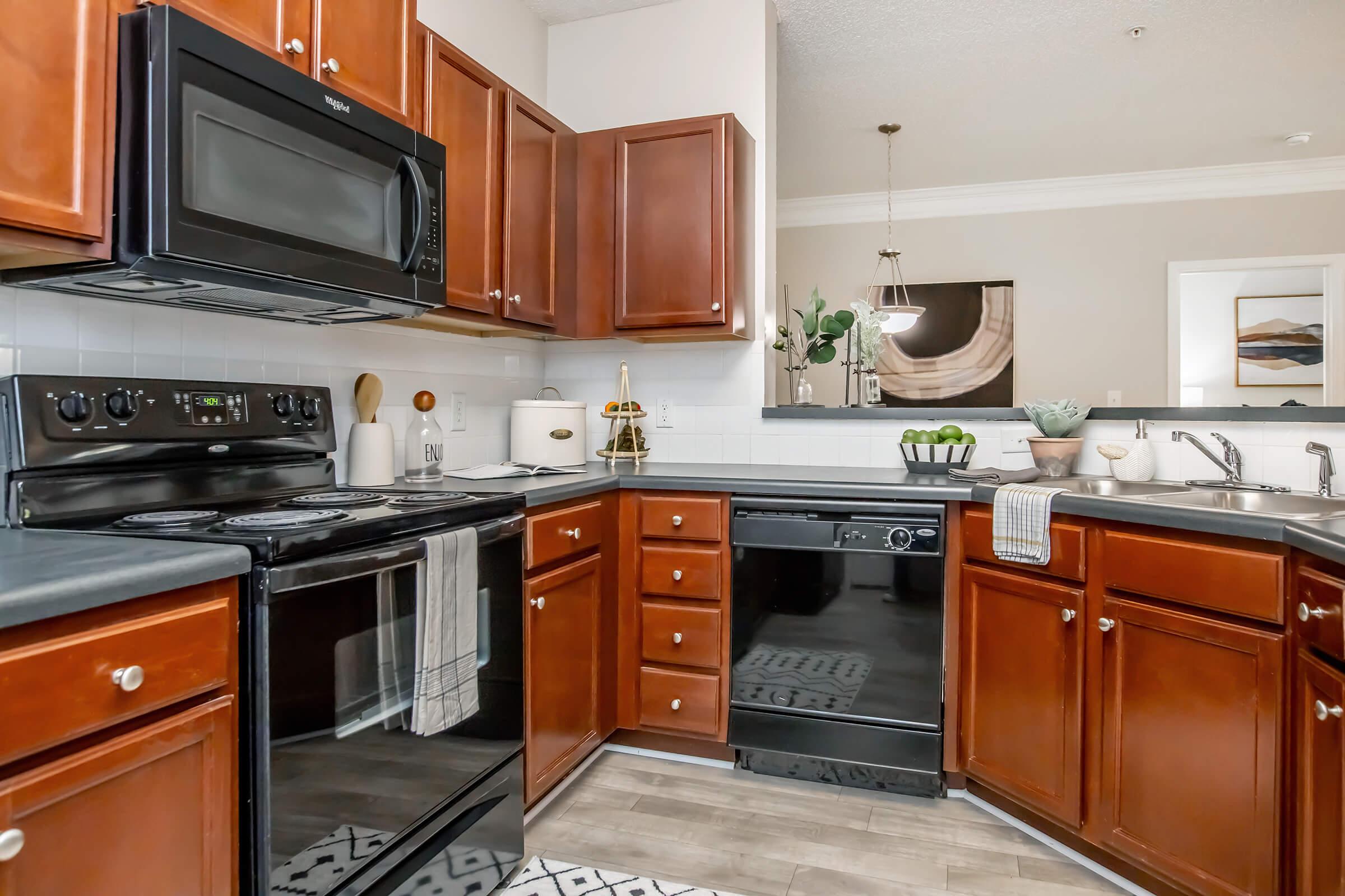 Modern kitchen featuring dark wood cabinets, black appliances including a microwave, oven, and dishwasher, white countertops, and a light-colored backsplash. The space is well-organized with decorative items like a plant and bowl of fruit, complemented by a stylish rug and light fixture above the dining area.