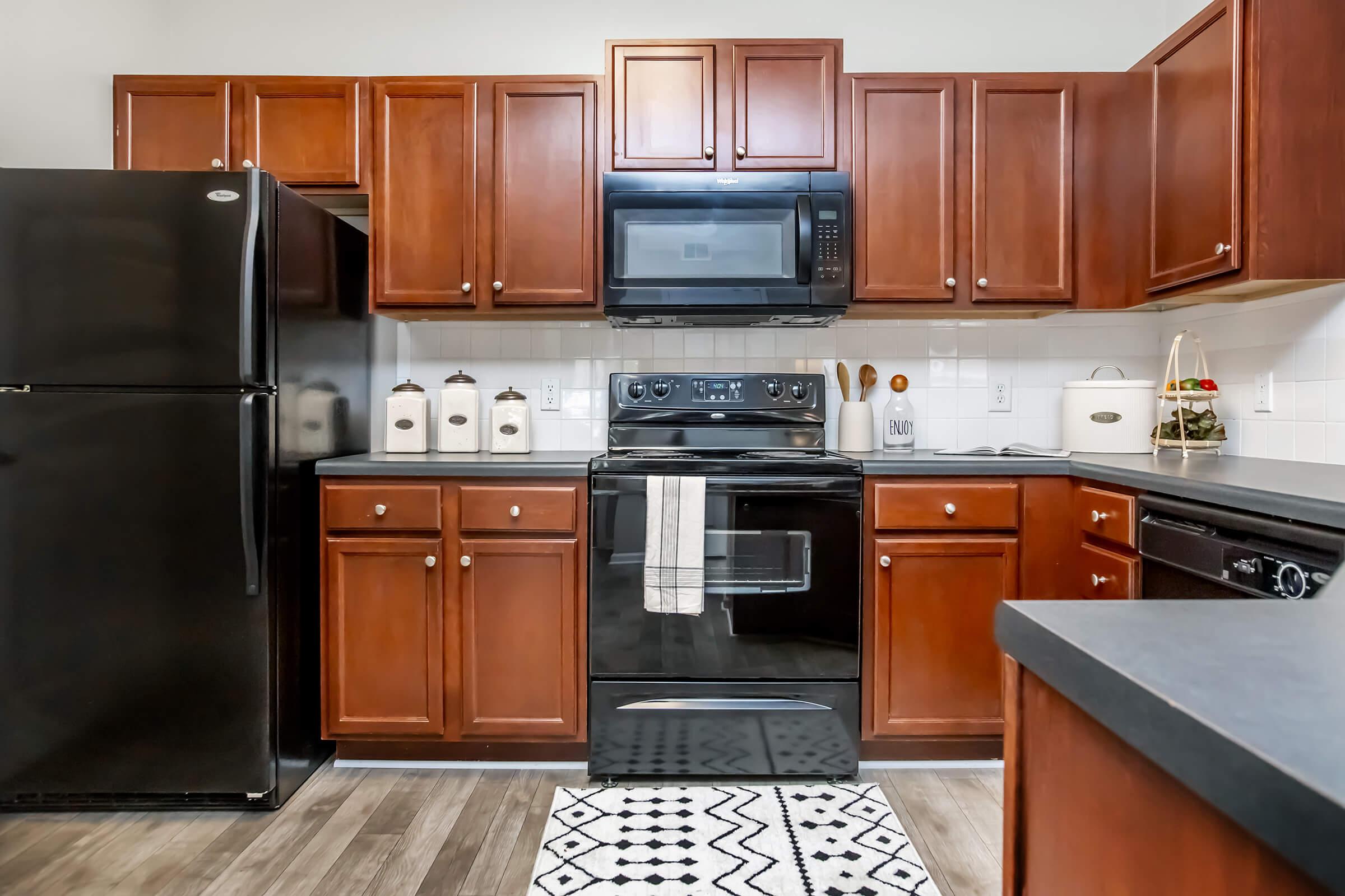 A modern kitchen featuring dark wood cabinets, a black refrigerator, and a black oven with a microwave above. The countertop is dark, and there is a patterned rug on the floor. A few decorative items, including canisters and a small plant, are placed on the counter. Natural lighting brightens the space.