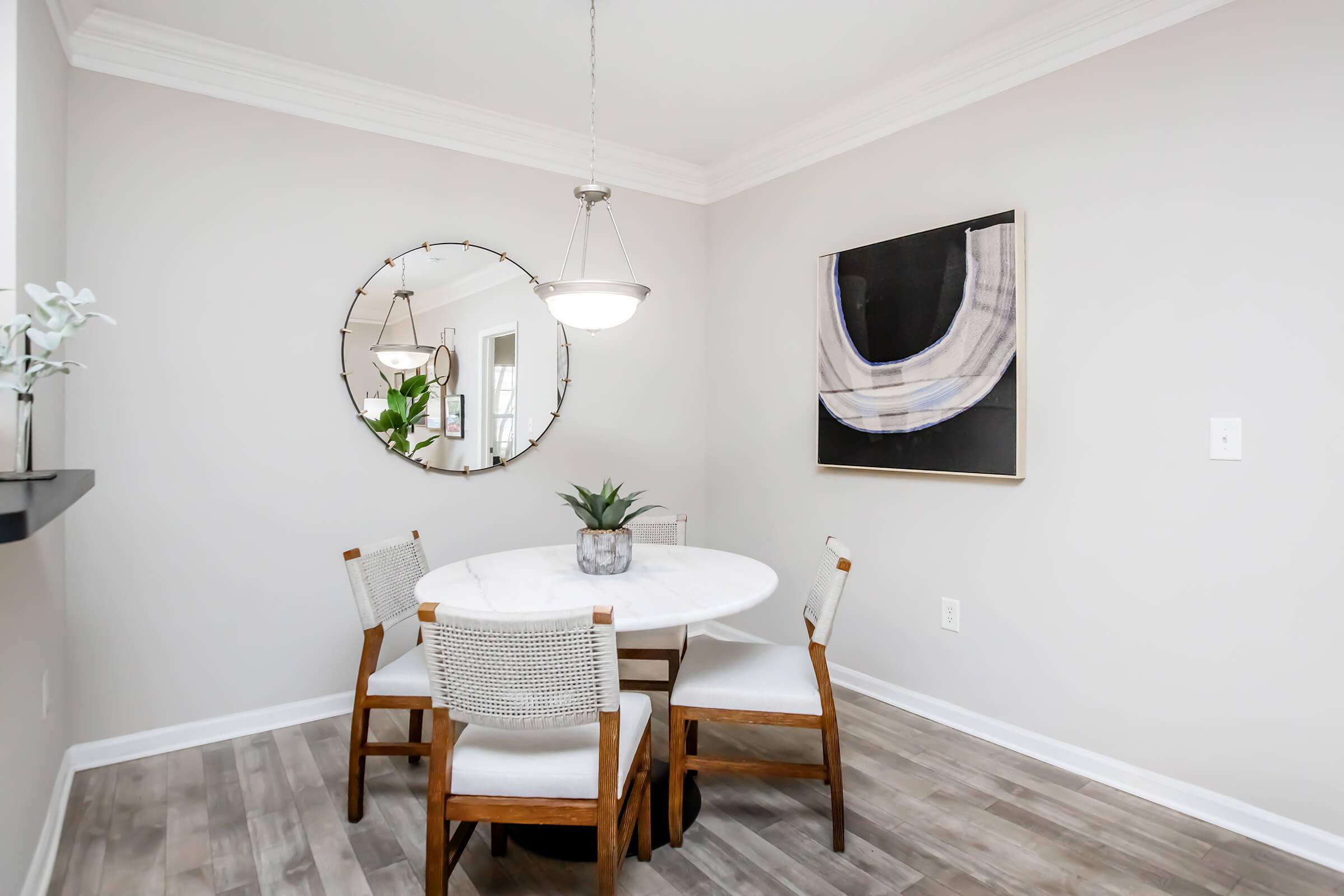 Cozy dining nook featuring a round marble table with four wooden chairs. A minimalist mirror hangs on the wall, reflecting light. A modern art piece is displayed above the table, and a small green plant adds a touch of greenery to the scene. The room has neutral gray walls and wooden flooring.