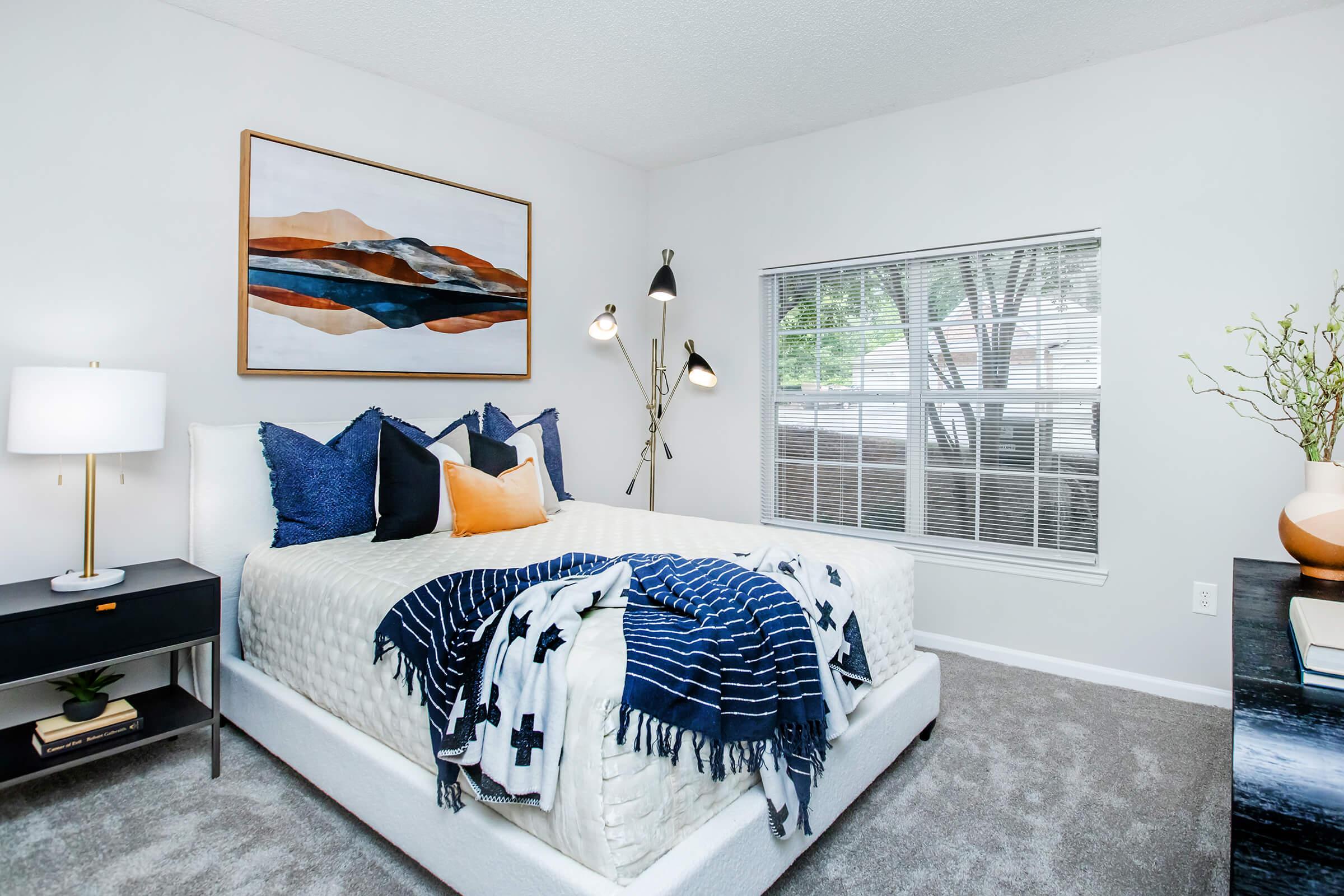 A cozy bedroom featuring a white bed adorned with a patterned blanket and colorful pillows. A modern floor lamp stands beside the bed, and a piece of abstract art hangs on the wall. There's a window with natural light coming in, and a bedside table with a lamp and some decor.