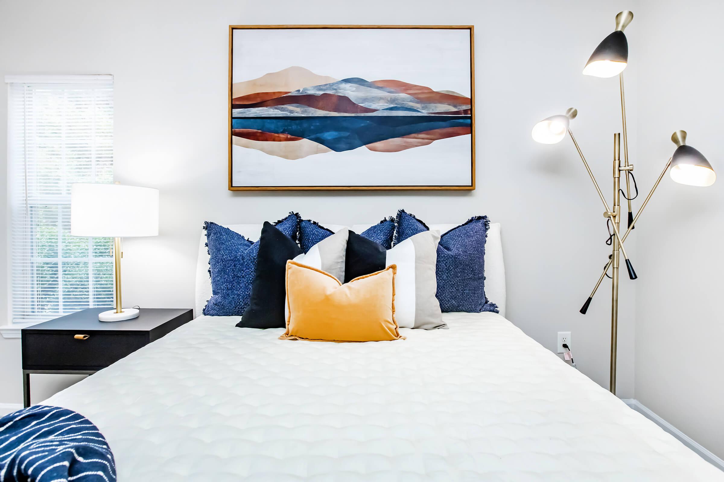 A cozy bedroom featuring a neatly made bed with decorative pillows, a bedside lamp, and a modern floor lamp. Above the bed is a large artwork depicting abstract mountains and a reflection in water. Natural light enters through a window, creating a warm atmosphere.