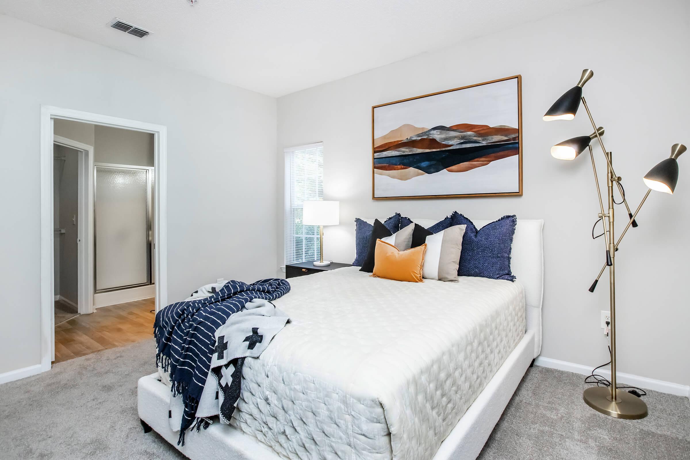 A cozy bedroom featuring a plush bed with decorative pillows, a modern wall art piece above, a stylish floor lamp, and a window allowing natural light. A bathroom door is visible in the background, with light-colored walls and soft carpeting creating a warm atmosphere.