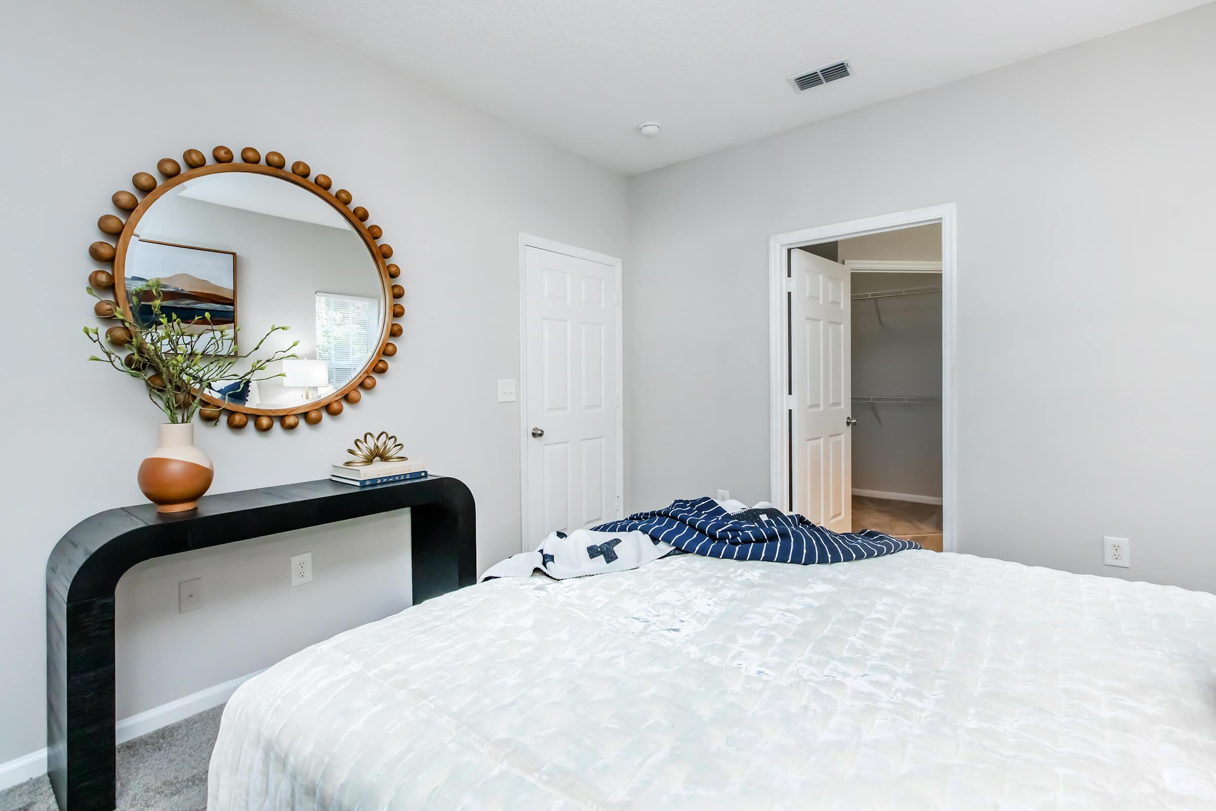 A cozy bedroom featuring a neatly made bed with crumpled bedding, a stylish mirror with a circular frame on the wall, a console table with decorative items, and a doorway leading to a closet. The walls are painted light gray, creating a calm atmosphere.