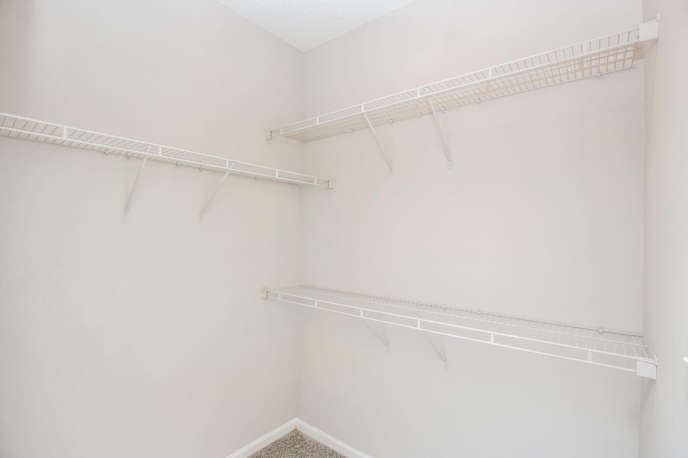 A spacious, empty closet with light gray walls and white wire shelving. The shelves are mounted on the walls, providing ample storage space. The floor is carpeted in a neutral tone, enhancing the clean, minimalist look of the closet.