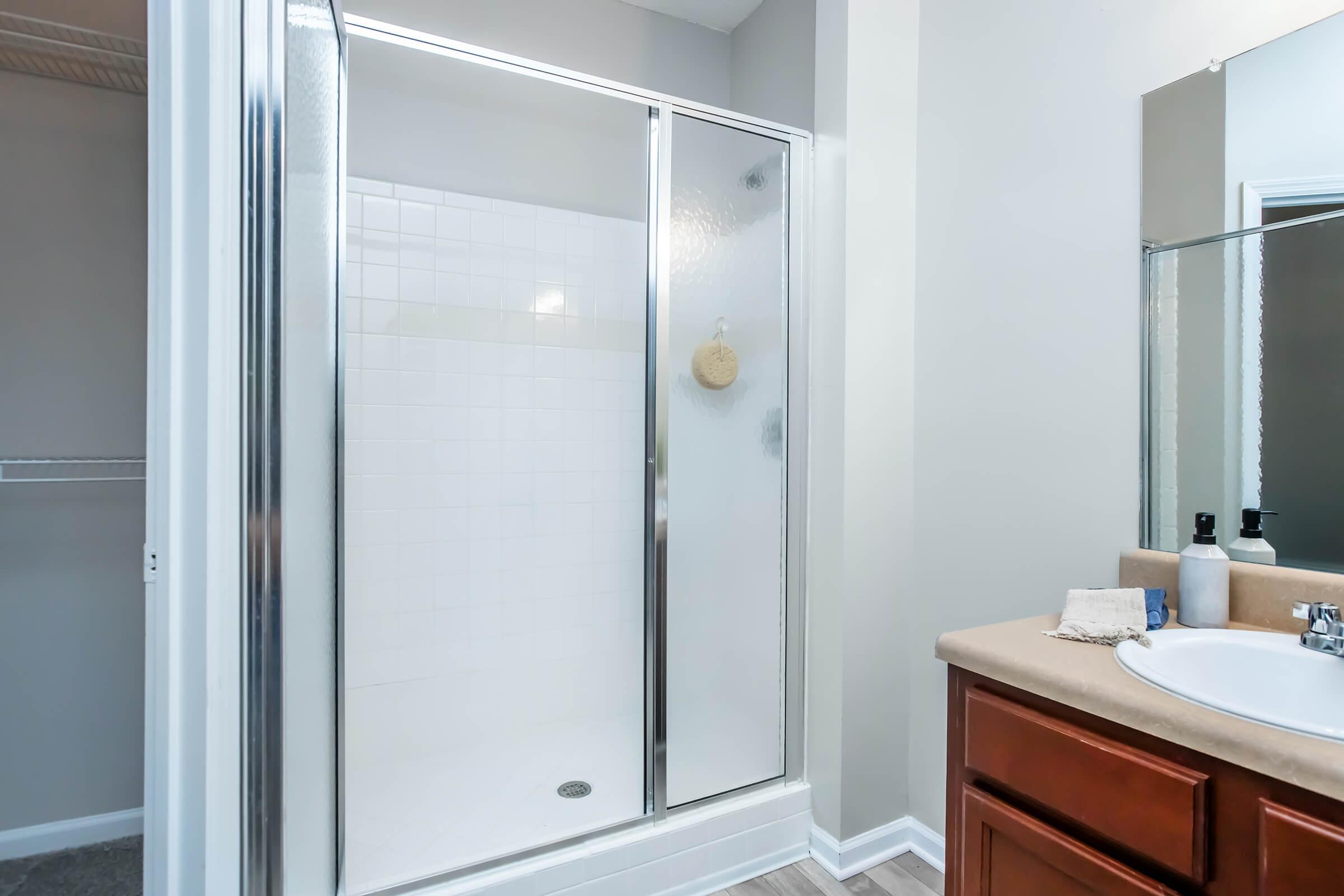 A clean bathroom featuring a glass shower enclosure with a tiled interior and a wall-mounted showerhead. There is a beige countertop with toiletries and a mirror, and visible light-colored walls and flooring. A closet space is partially seen on the left.