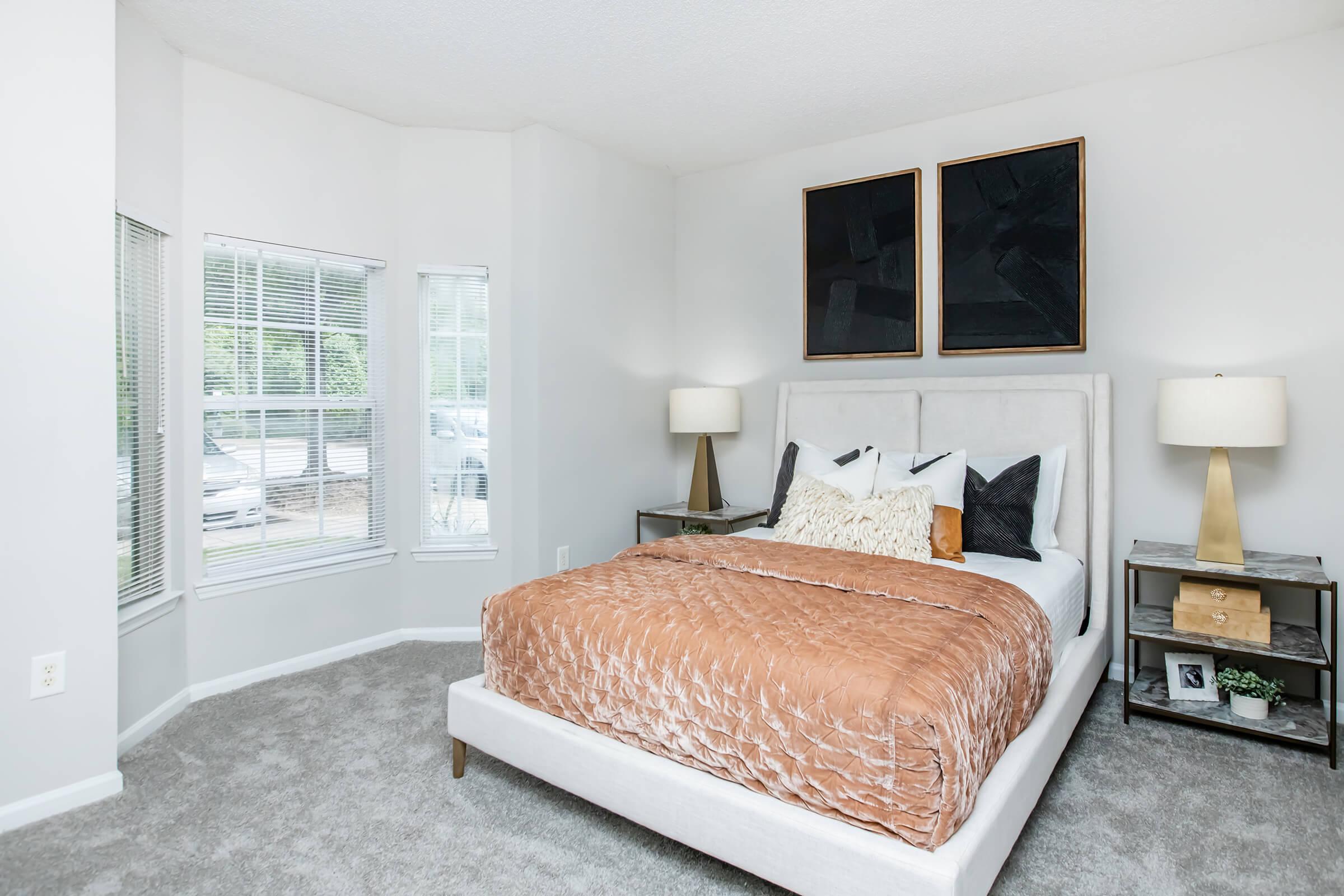 Cozy bedroom featuring a light-colored bed with an orange-brown quilt and decorative pillows. Two abstract paintings hang above the headboard. Beside the bed, there are stylish nightstands with lamps, and large windows allow natural light to fill the space, enhancing the inviting atmosphere. Soft gray carpeting completes the room.