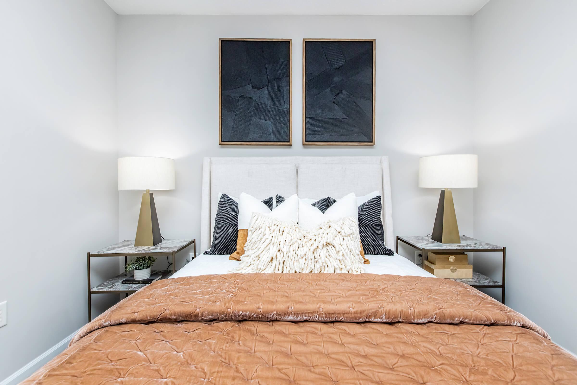 A cozy bedroom featuring a white bed with an orange-brown quilt and decorative pillows. On either side of the bed, there are modern bedside tables with stylish lamps. Above the bed, two framed black wall art pieces add a contemporary touch. The walls are painted light gray, creating a calm atmosphere.
