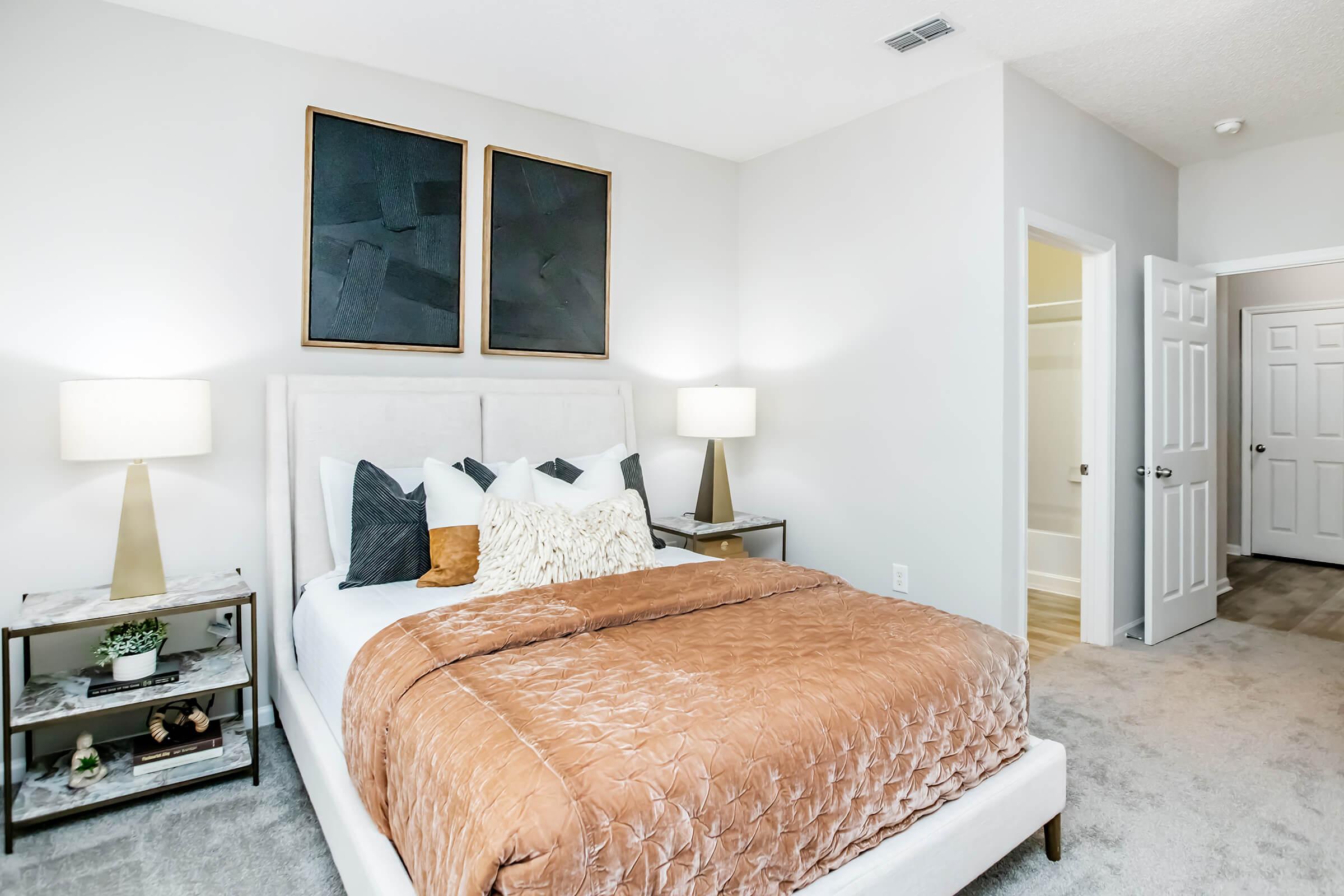 A modern bedroom featuring a light-colored bed with decorative pillows, an elegant brown quilt, and two stylish nightstands on either side with lamps. The walls are painted in a soft gray, and there are two abstract art pieces above the bed. A bathroom door is visible in the background.
