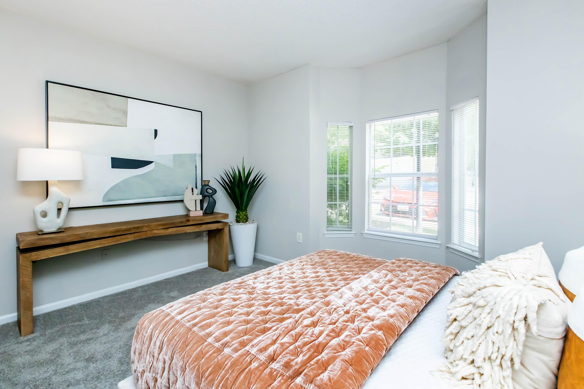 A cozy bedroom featuring a soft brown quilted bedspread, a decorative pillow, and a contemporary artwork on the wall. There is a wooden console table with a lamp and decorative items, along with a potted plant in a white planter and large windows letting in natural light. The walls are painted in a light gray tone.