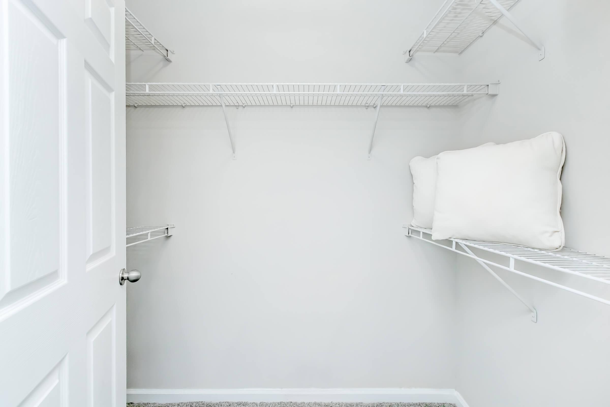 An empty closet with white wire shelving on the walls. A white pillow is resting on one of the shelves, while the walls and floor are painted in light colors, creating a bright and spacious appearance. The closet door is closed, showing a simple design.
