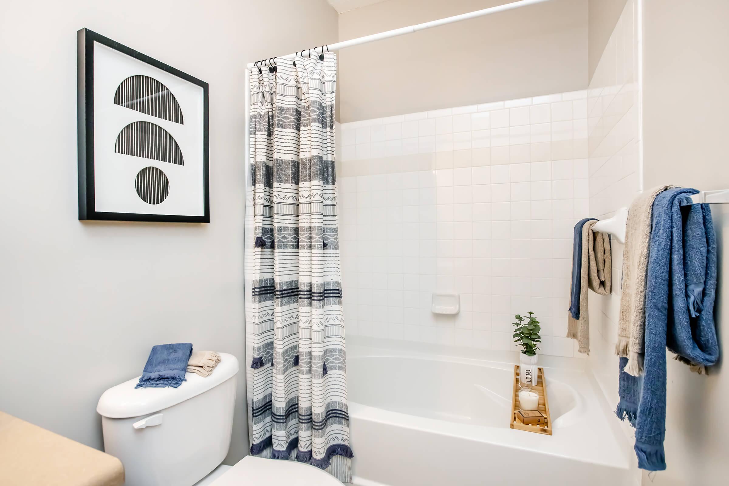 A modern bathroom featuring a bathtub with a white shower curtain, a decorative wall art piece, a toilet, and towels hanging on a rack. A small plant is placed on a wooden stand beside the bathtub, creating a clean and inviting atmosphere.