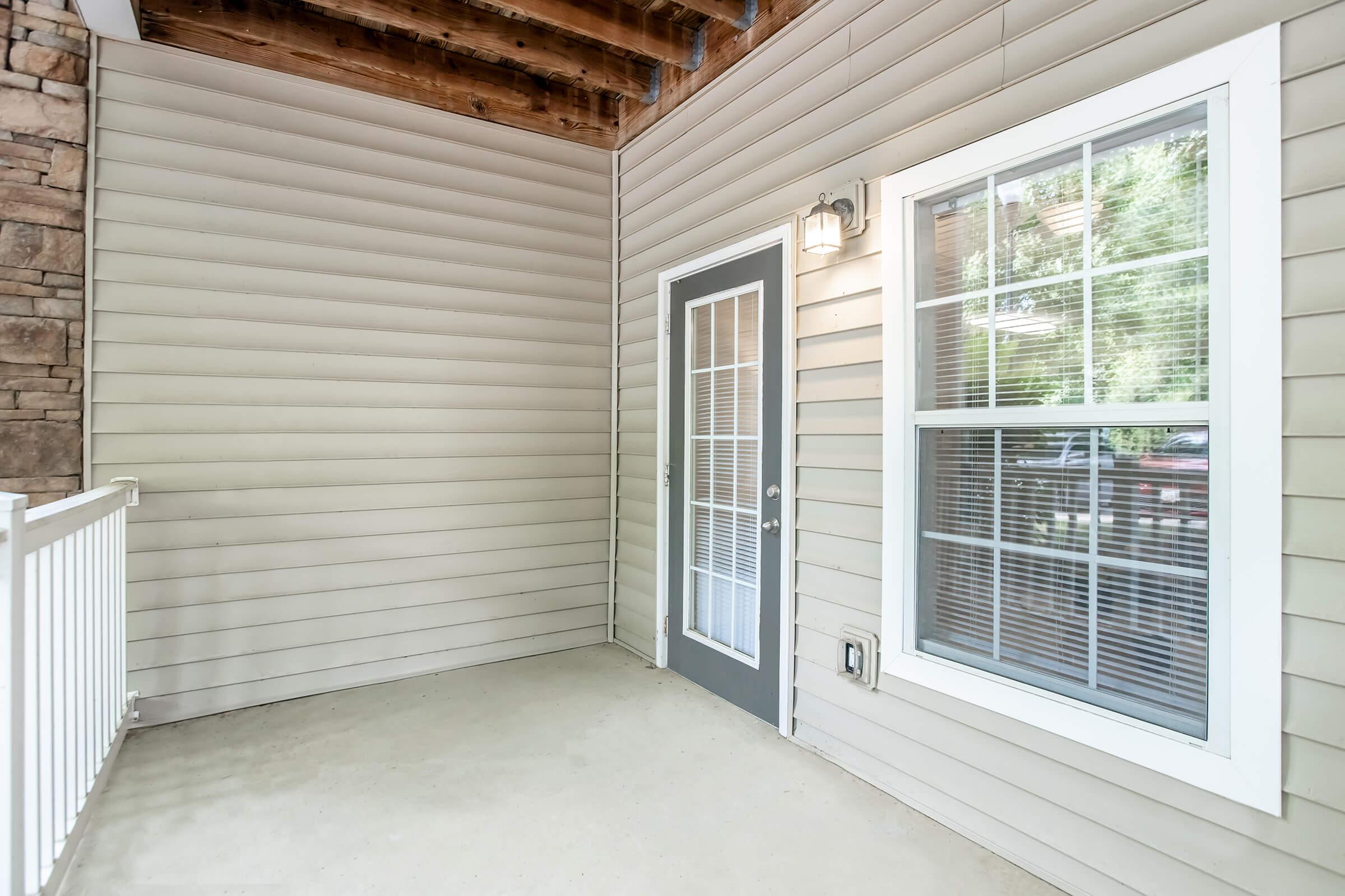 A small outdoor porch area with a light-colored floor, beige vertical siding, and a gray door with a glass insert. There's a window with white framing adjacent to the door. The ceiling is supported by wooden beams, creating a cozy and inviting space.