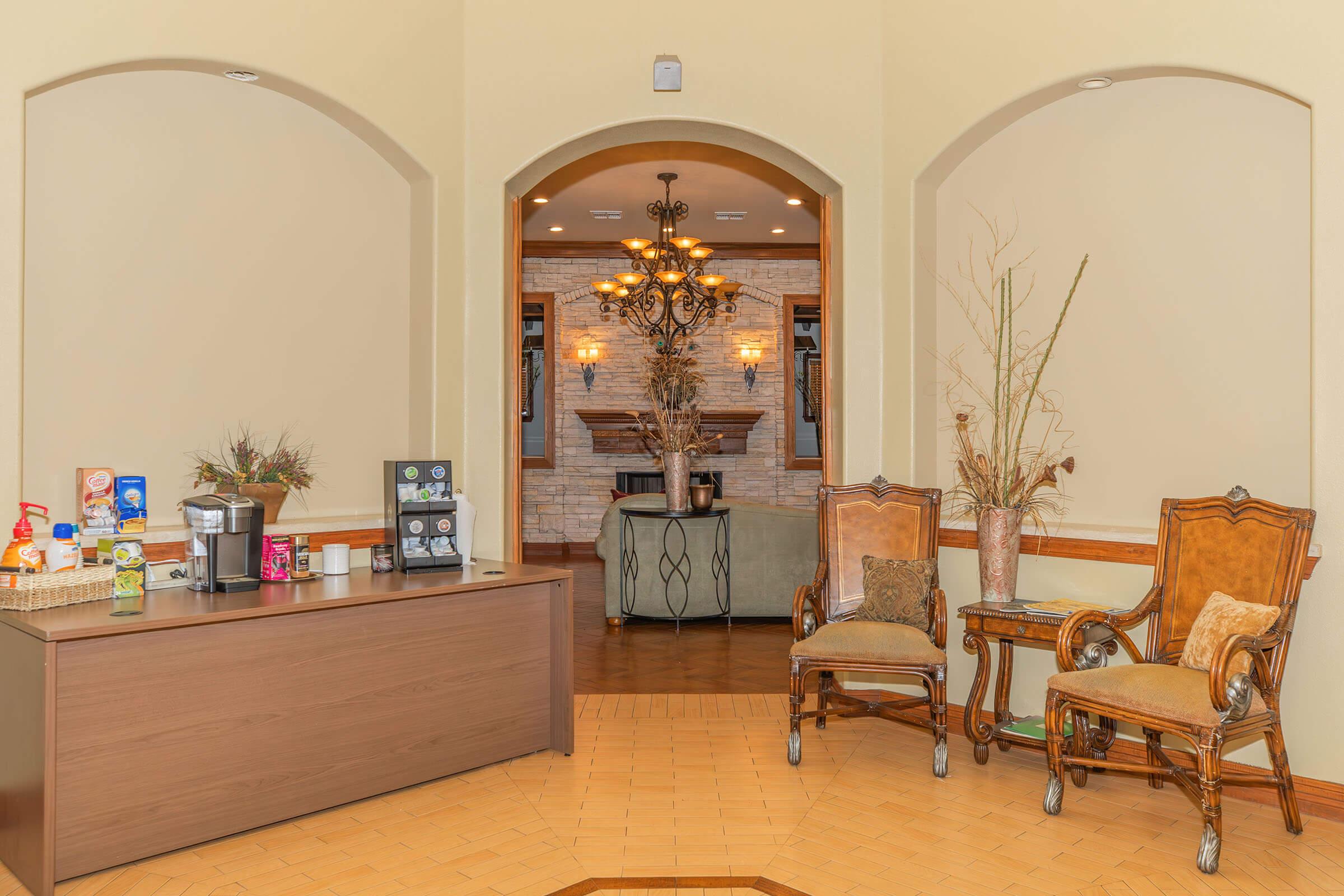 Interior view of a cozy waiting area featuring a wooden reception desk with various beverages and snacks. Two elegant chairs are positioned next to the desk, and a doorway leads to a well-lit room with a chandelier and a fireplace in the background. Decorative elements add warmth to the space.