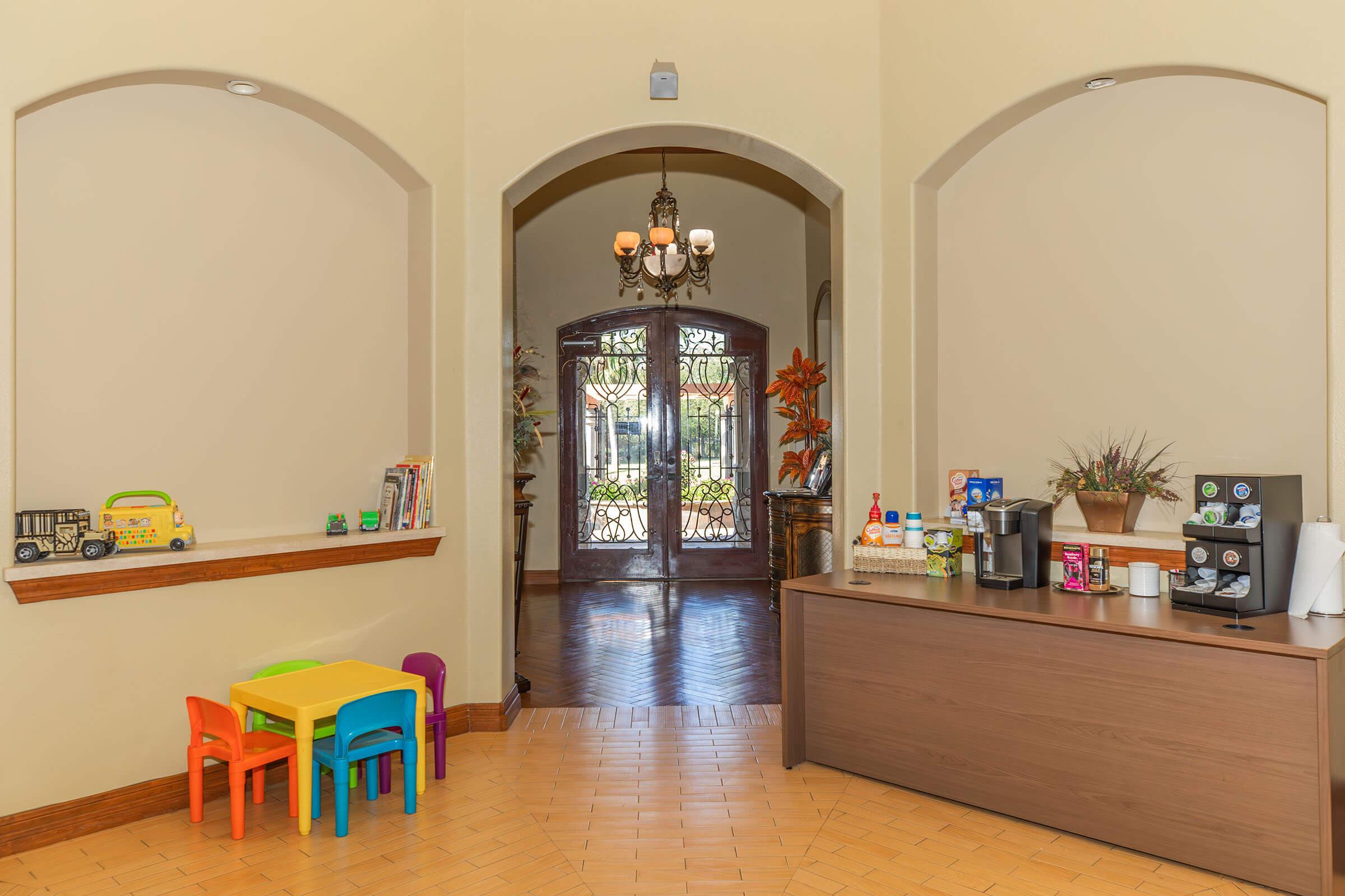 Interior view of a welcoming hallway leading to double doors with decorative ironwork. To the right, a coffee station with various beverage options and condiments. To the left, a small children's table in bright colors. The space features warm lighting and stylish decor, creating an inviting atmosphere.