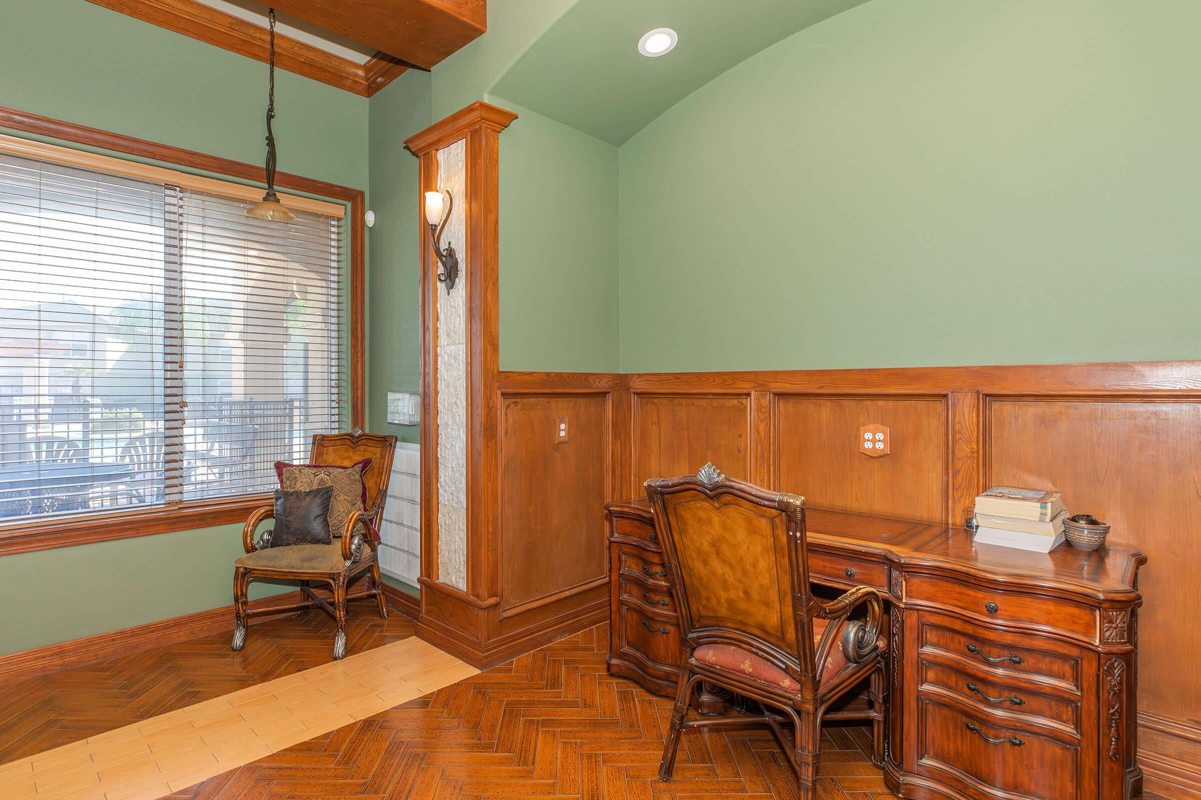 A cozy home office with green walls and polished wood accents. It features a vintage wooden desk with books and a small decorative bowl on top. To the left, a classic upholstered chair sits next to a large window with blinds, providing natural light. The floor has a herringbone pattern, adding warmth to the space.
