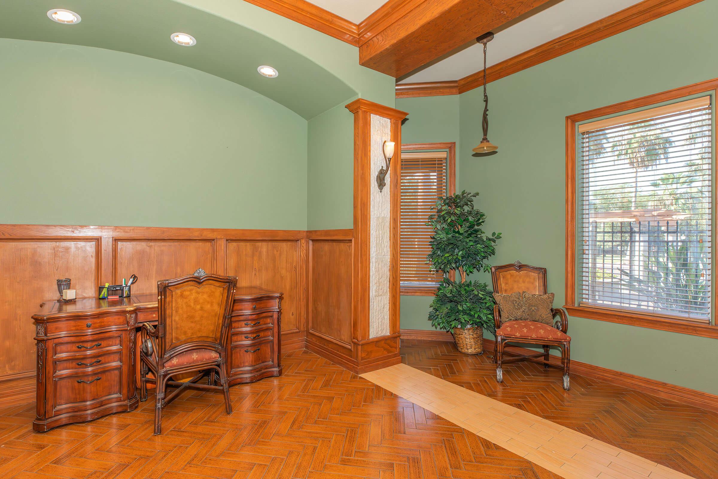 A well-lit office space featuring a wooden desk with a chair, two ornate chairs, and a potted plant. The walls are painted green with wood paneling, and there are large windows with blinds allowing natural light. The floor consists of herringbone-patterned wood, creating a warm and inviting atmosphere.
