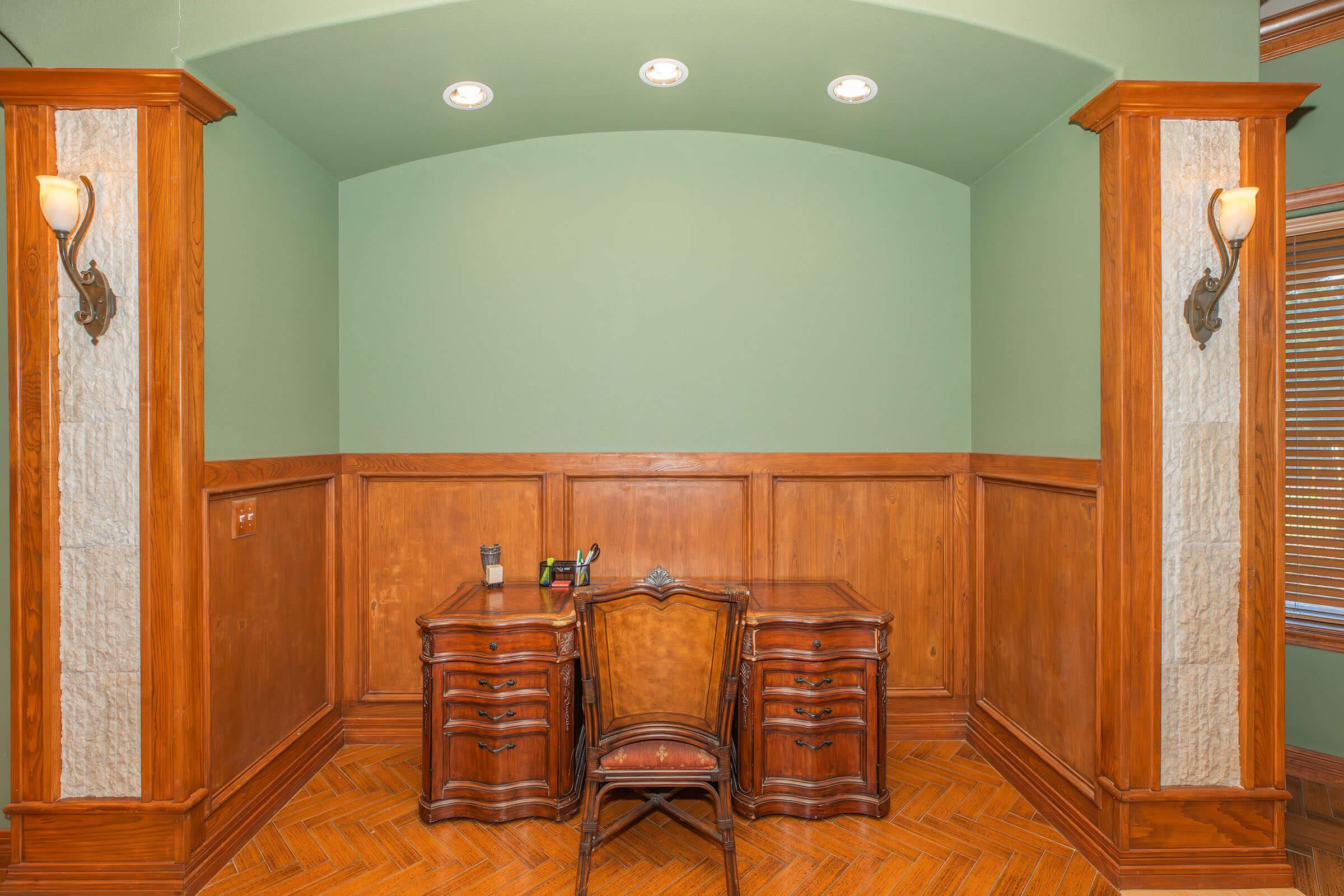 A cozy home office featuring a traditional wooden desk with ornate detailing, flanked by two decorative wall sconces. The walls are painted in a soft green, complementing the rich wooden paneling. The herringbone wood floor adds warmth to the space, creating an inviting atmosphere for work or reading.