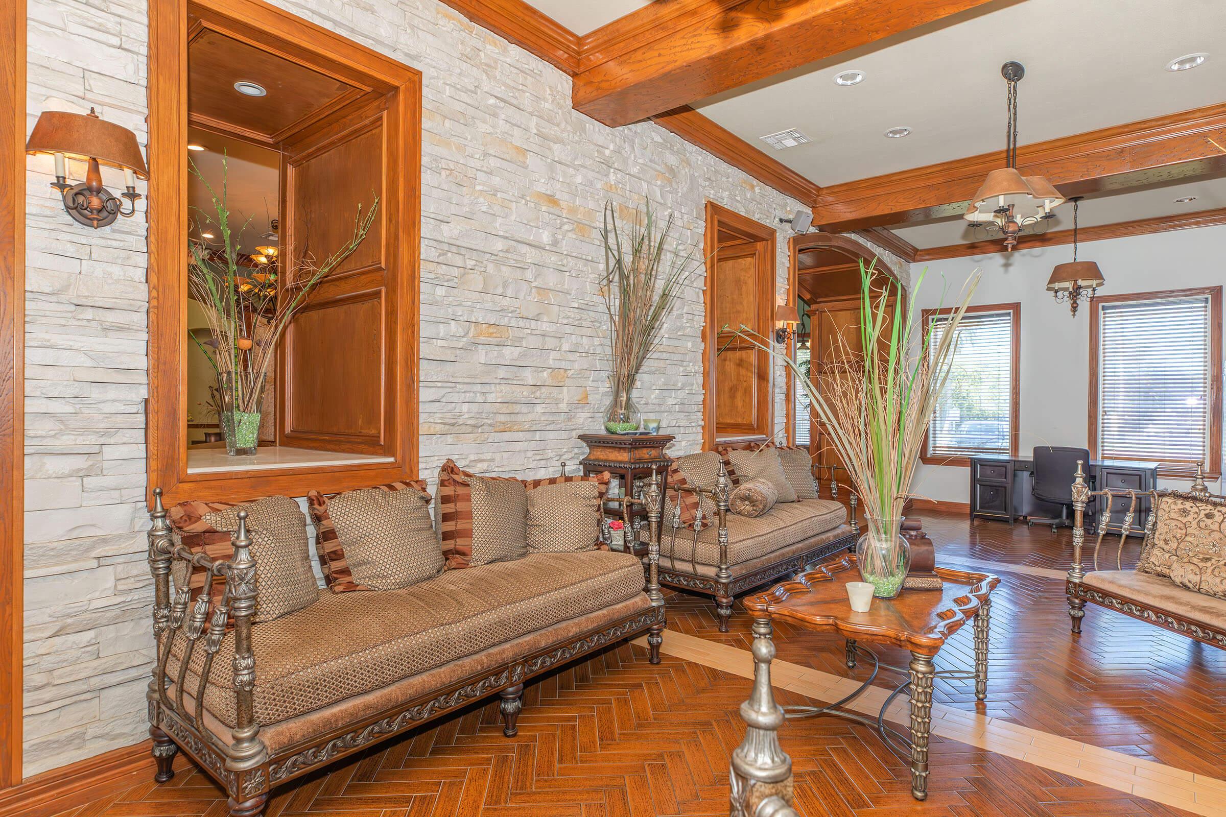 Elegant living room featuring two wooden sofas adorned with intricate carvings and soft cushions, complemented by a wooden coffee table. The walls are accented with stone panels, and there are decorative plants in vases. Ample natural light filters through windows with wooden frames, creating a warm and inviting atmosphere.