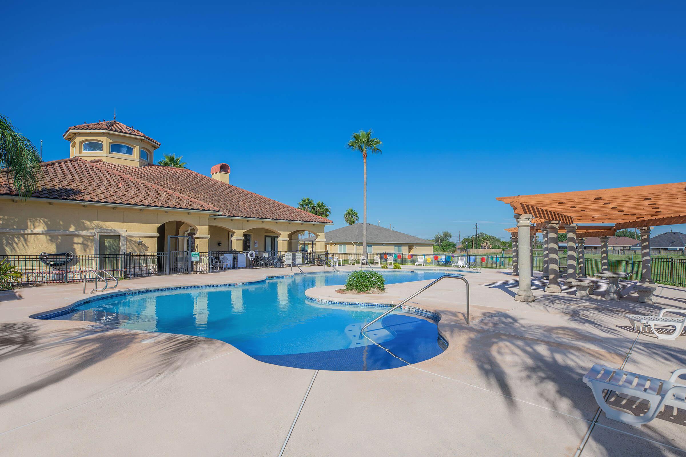 A bright, sunny pool area featuring a blue swimming pool surrounded by lounge chairs. In the background, there is a clubhouse with a tiled roof and a pergola offering shaded seating. Palm trees line the poolside, and a clear blue sky completes the serene atmosphere.