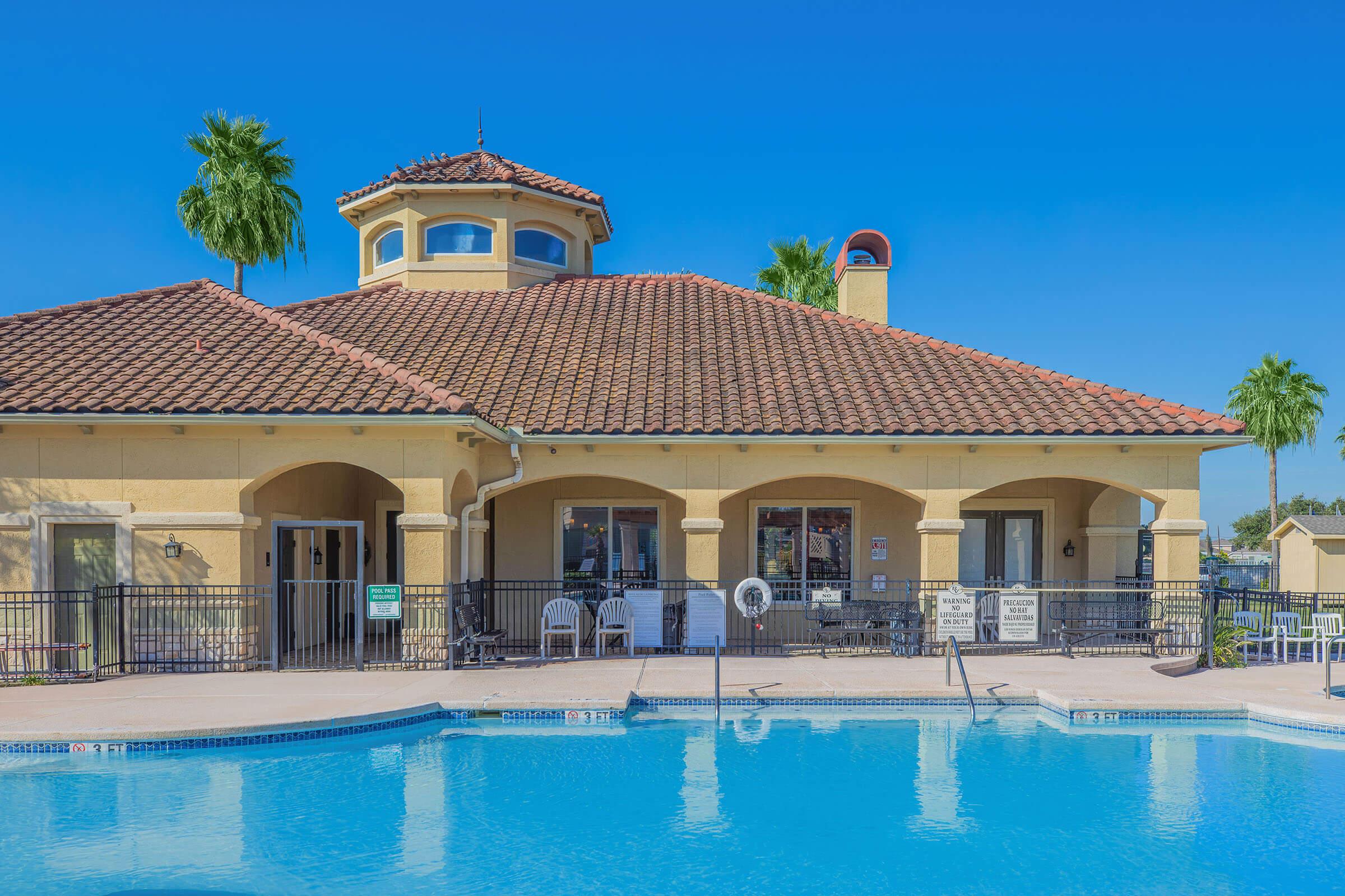 A picturesque swimming pool area featuring a large, well-maintained pool in front of a two-story building with a red-tiled roof and arched entryways. The scene is complemented by tall palm trees and a clear blue sky, creating a relaxed and inviting atmosphere.