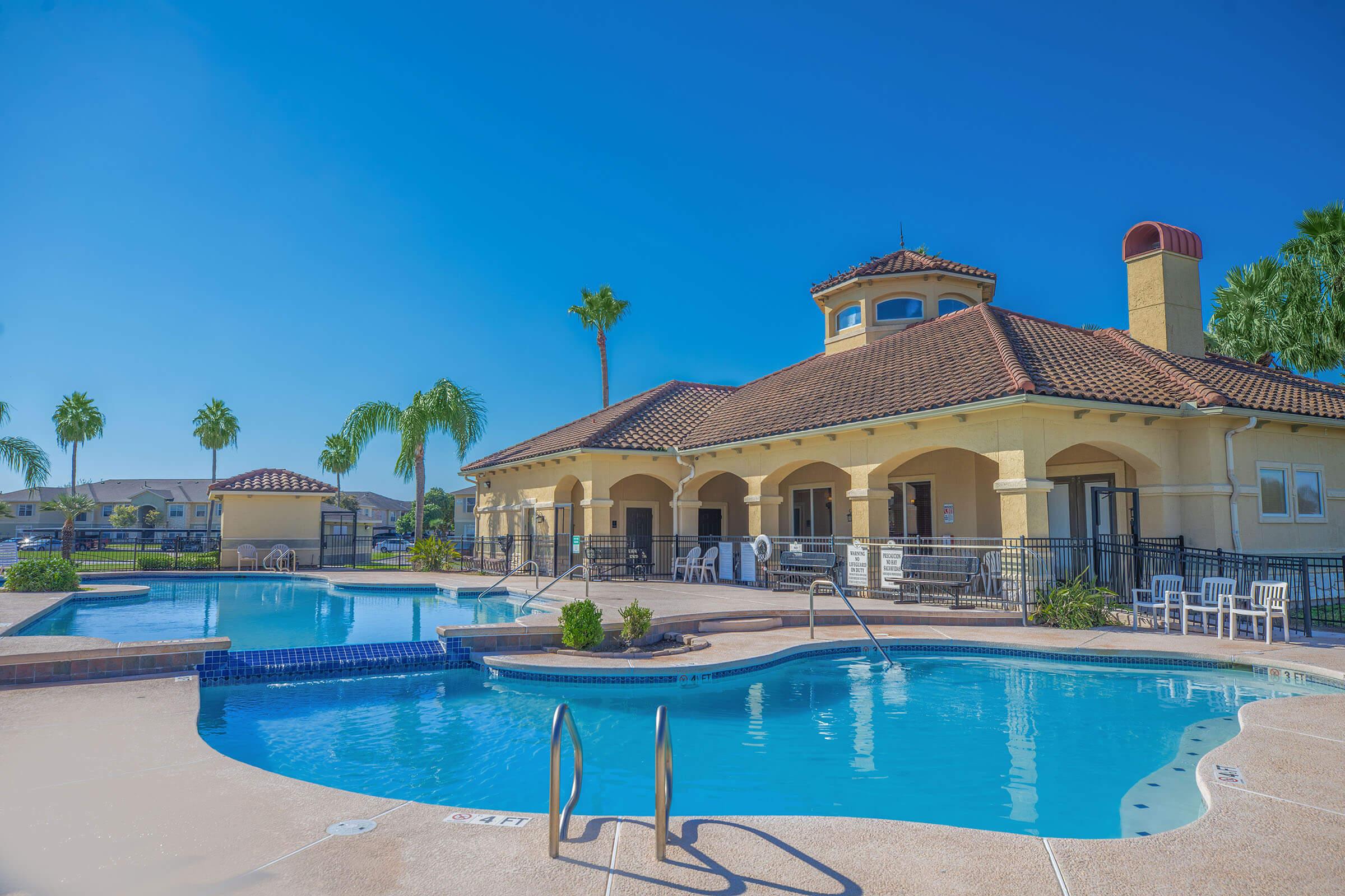 A sunny pool area featuring two swimming pools with clear blue water, surrounded by palm trees. There is a clubhouse with a tiled roof in the background. Several white lounge chairs are positioned around the pool deck, and a bright blue sky complements the vibrant scene.