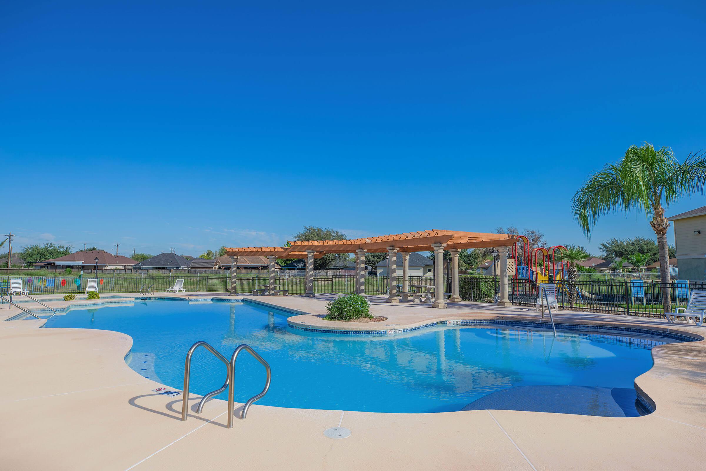 A clear blue swimming pool surrounded by a patio. There's a wooden pergola providing shade, with palm trees nearby. In the background, there are residential buildings and colorful playground equipment. The sky is bright blue with a few scattered clouds.