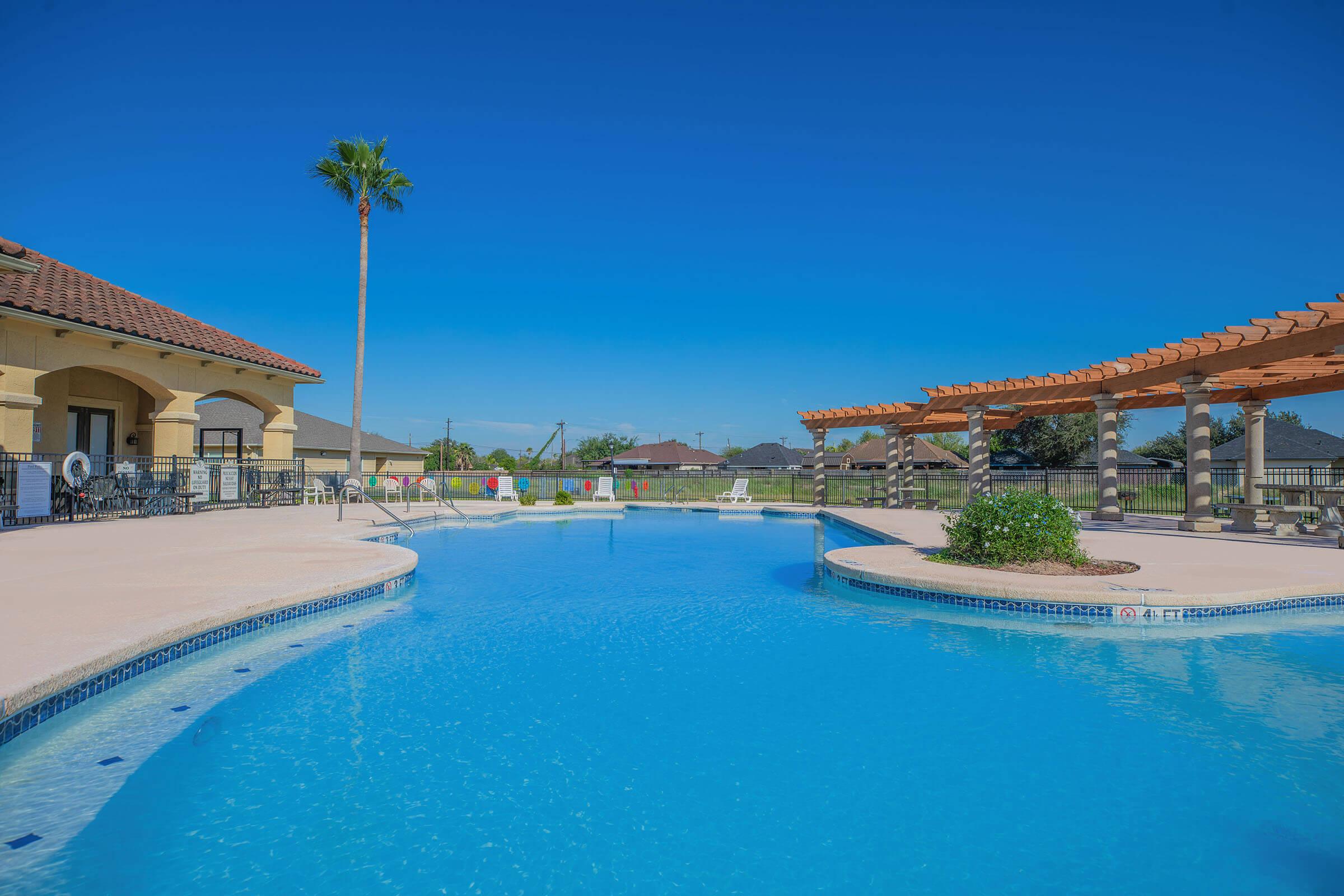 A clear blue swimming pool surrounded by a tan deck, lounge chairs, and palm trees. In the background, there are houses and a pergola. The sky is bright blue and cloudless, suggesting a sunny day.