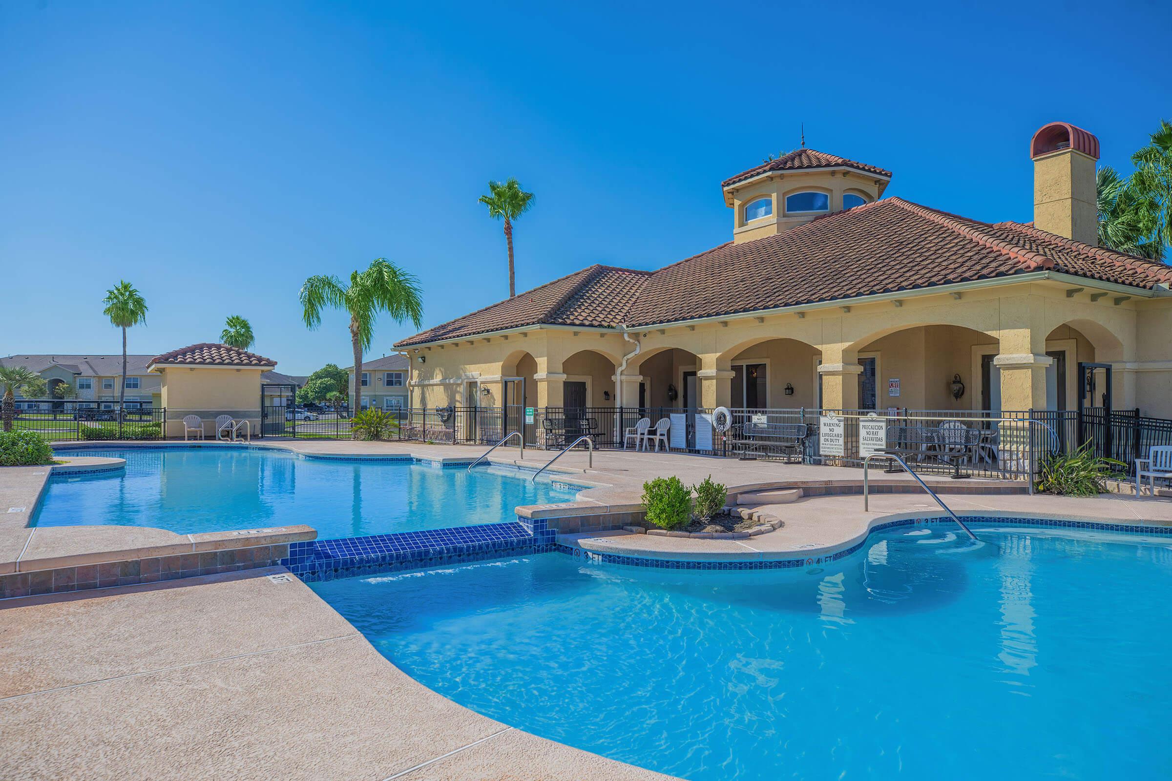 A bright blue swimming pool with a shallow end and steps, surrounded by palm trees. Adjacent is a yellow building with a red-tiled roof, featuring large arched windows and a patio area. The clear sky and vibrant colors create a warm, inviting atmosphere.