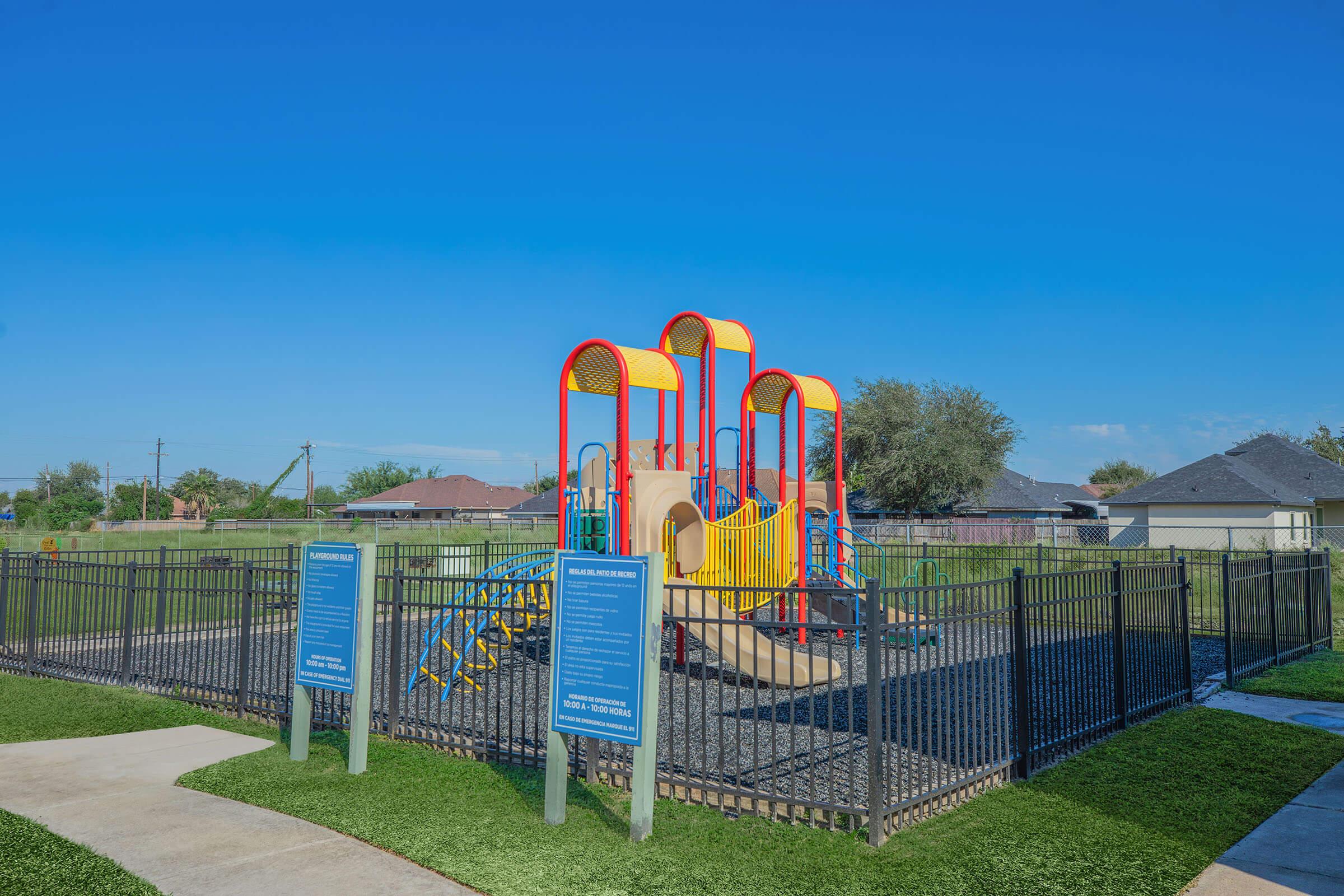 A colorful playground structure featuring slides and climbing areas, surrounded by a black fence. Signs near the entrance provide information about park rules. The playground is located in a grassy area under a clear blue sky, with residential homes visible in the background.