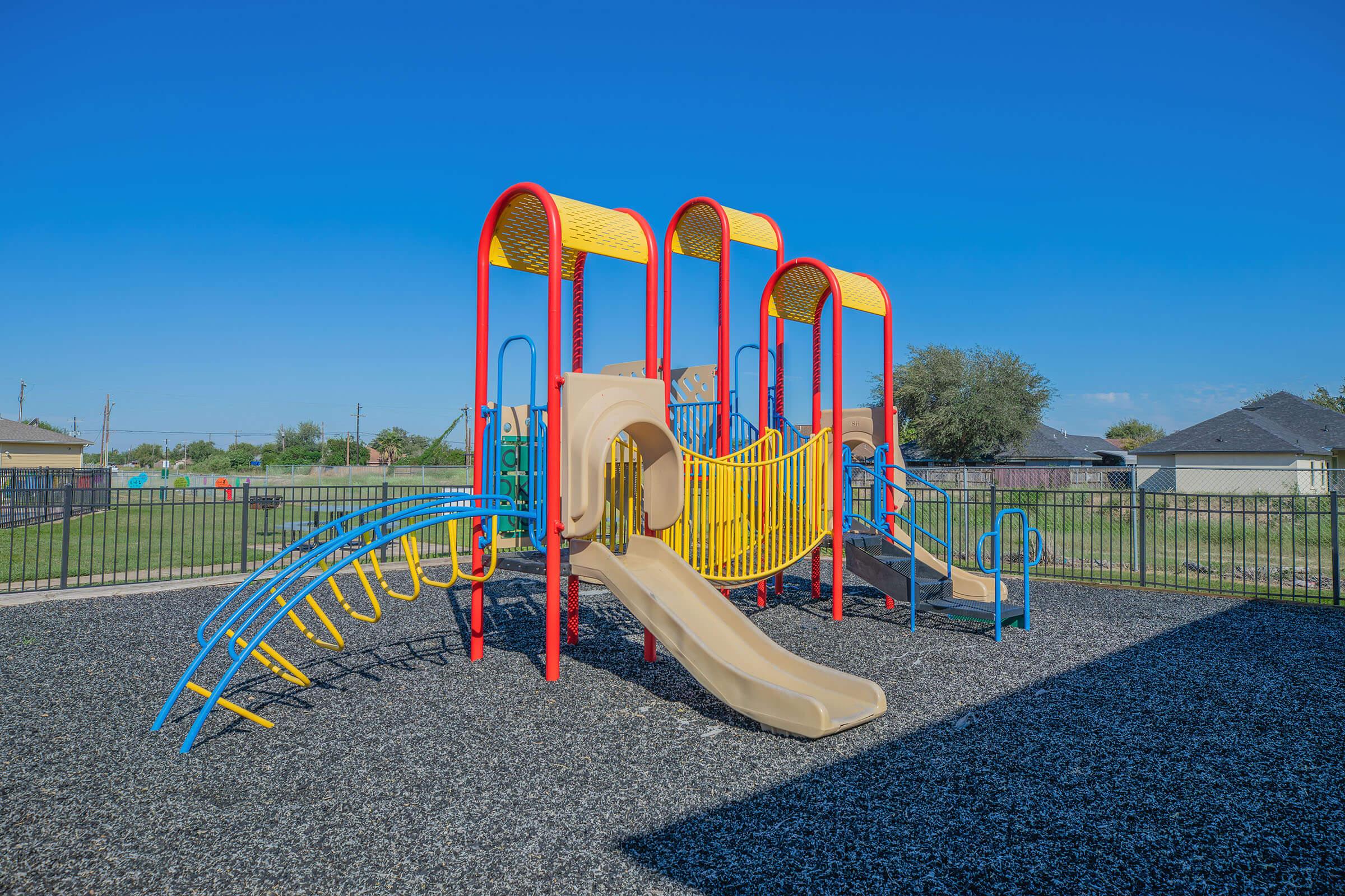 Colorful playground structure with multiple slides and climbing features, surrounded by a grassy area and a black rubber surface. Clear blue sky in the background, with residential homes visible in the distance.