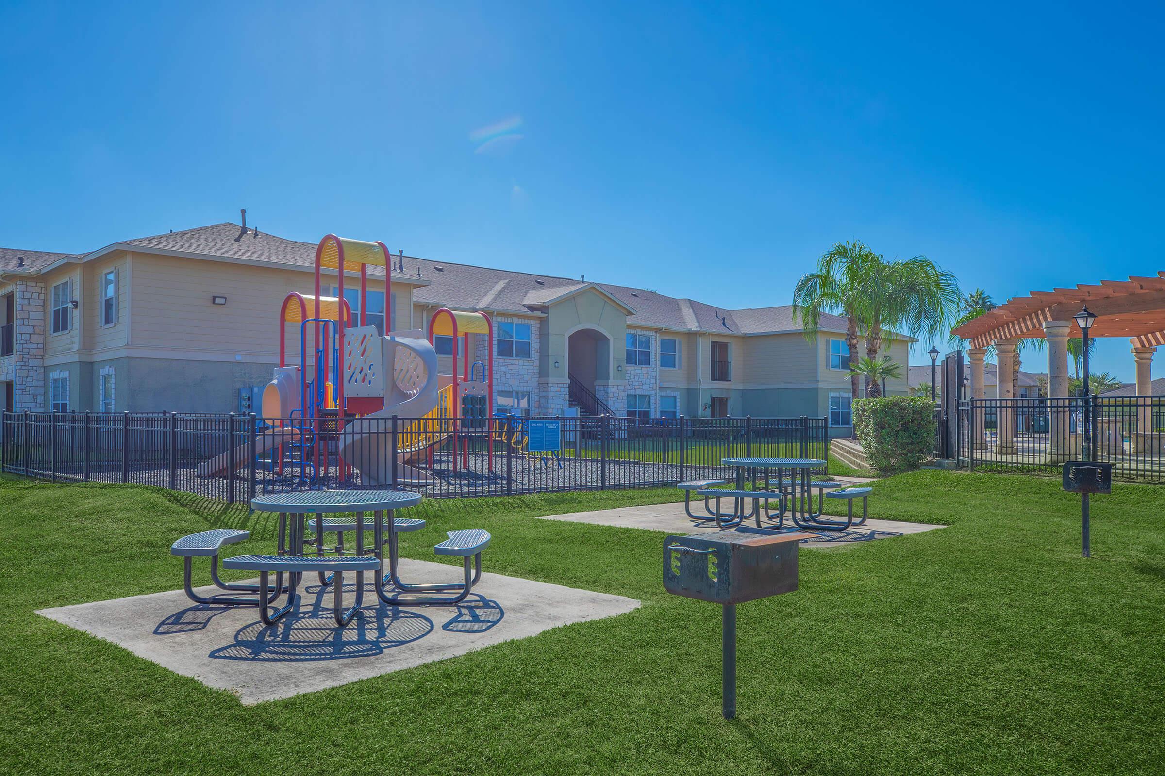 A sunny playground area featuring colorful play structures, picnic tables, and barbecue grills on a well-maintained lawn. In the background, residential buildings are visible, surrounded by a fence and palm trees, creating a welcoming outdoor space for families and children.