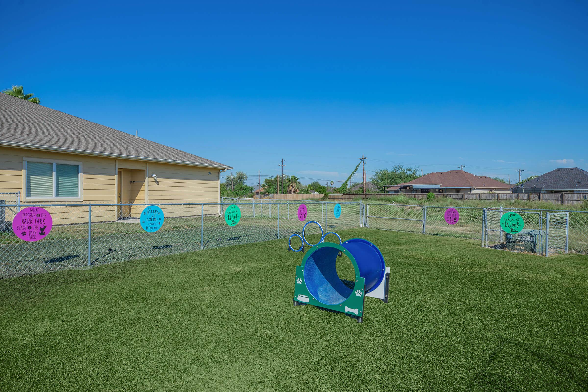 A fenced outdoor area featuring green grass, a blue tunnel and circular agility equipment for pets. Brightly colored signs adorn the fence, promoting positive messages. In the background, a single-story yellow house is visible under a clear blue sky.