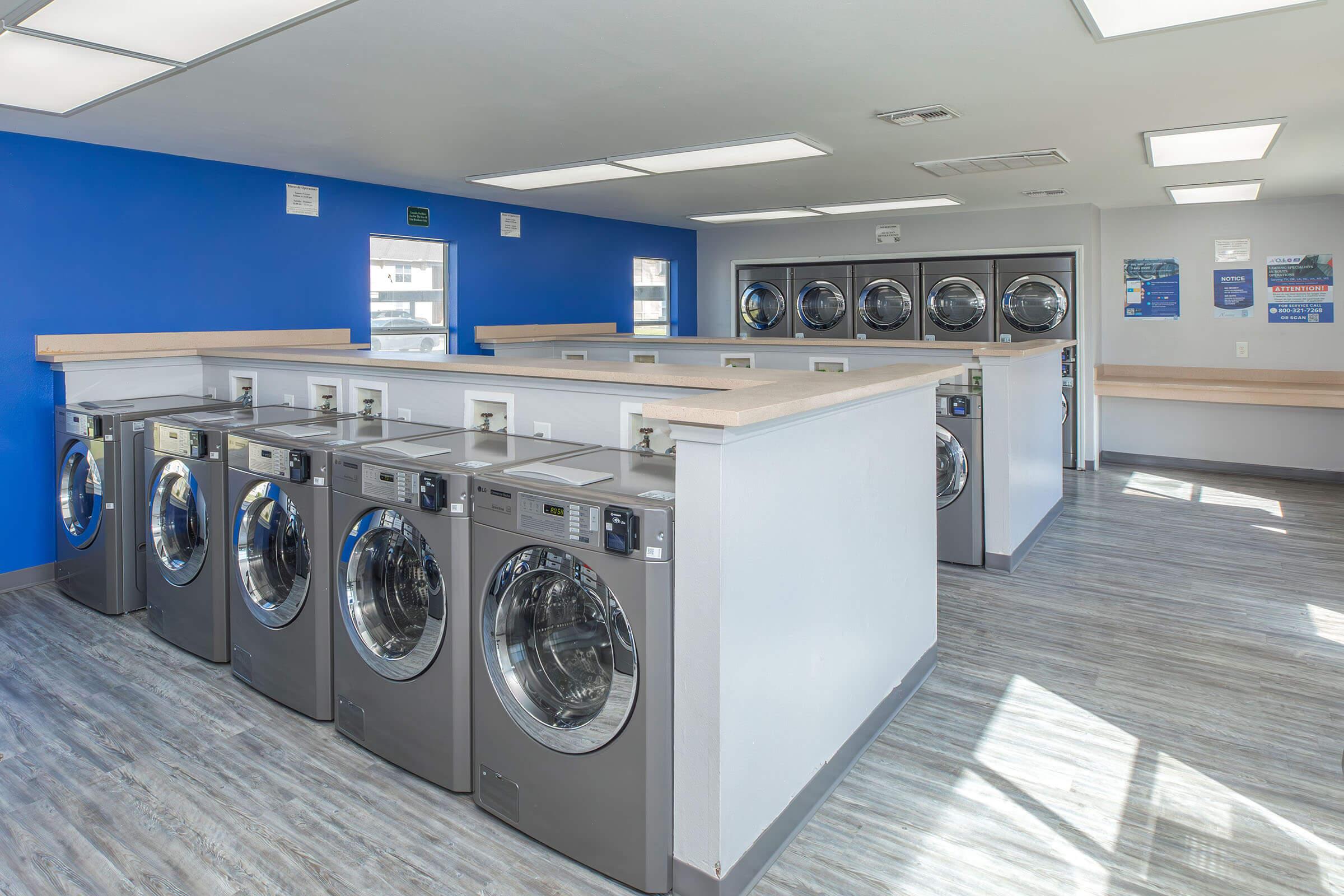 A bright and clean laundromat interior featuring several silver washing machines lined up against a wall. The walls are painted blue and light gray, with large windows allowing natural light. There are folding tables and an organized layout, creating a welcoming atmosphere for customers.
