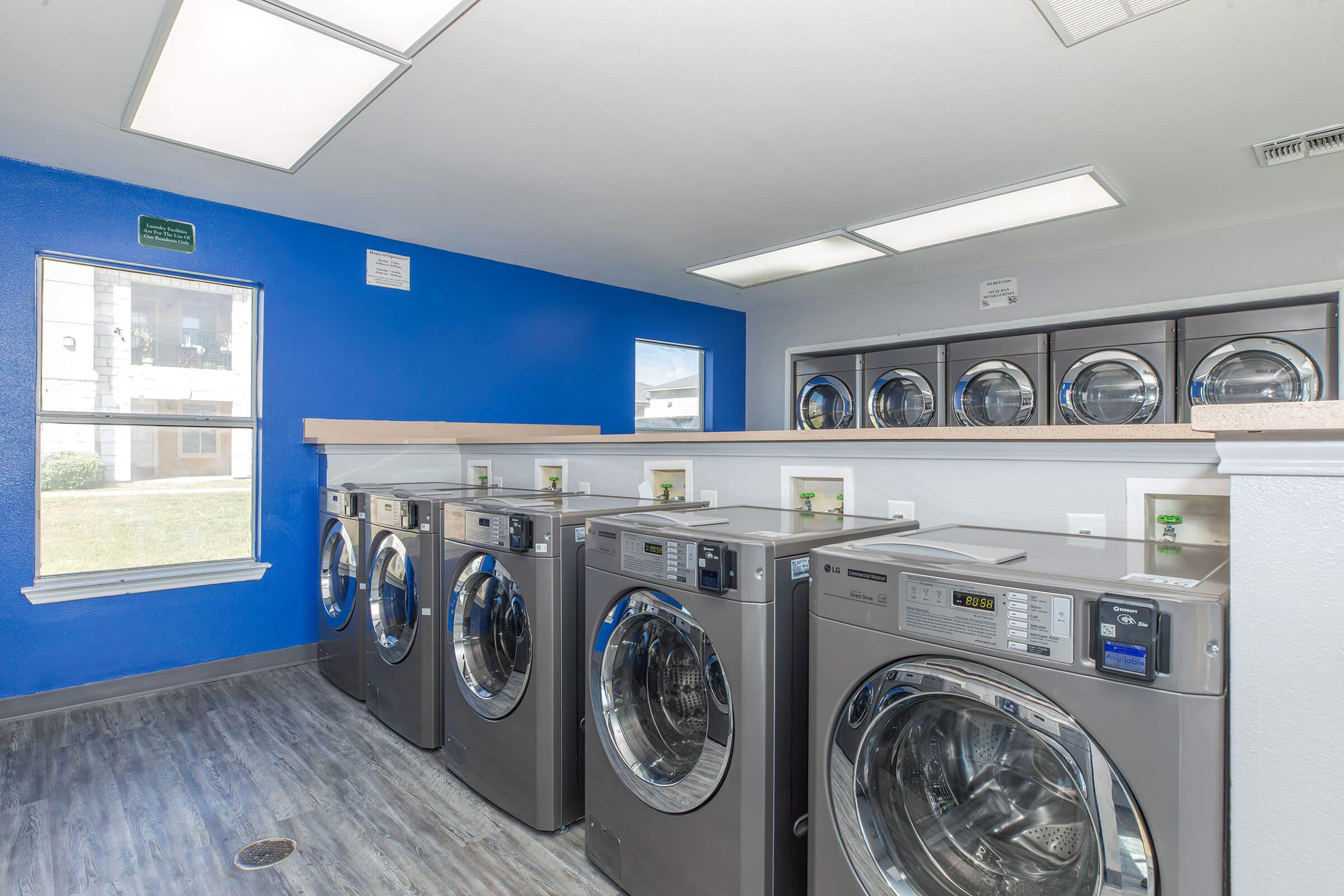 A modern laundry room featuring multiple stainless steel washing machines and dryers lined up against a bright blue wall. There are large windows allowing natural light, with a countertop space above the machines and a clean, tidy atmosphere.