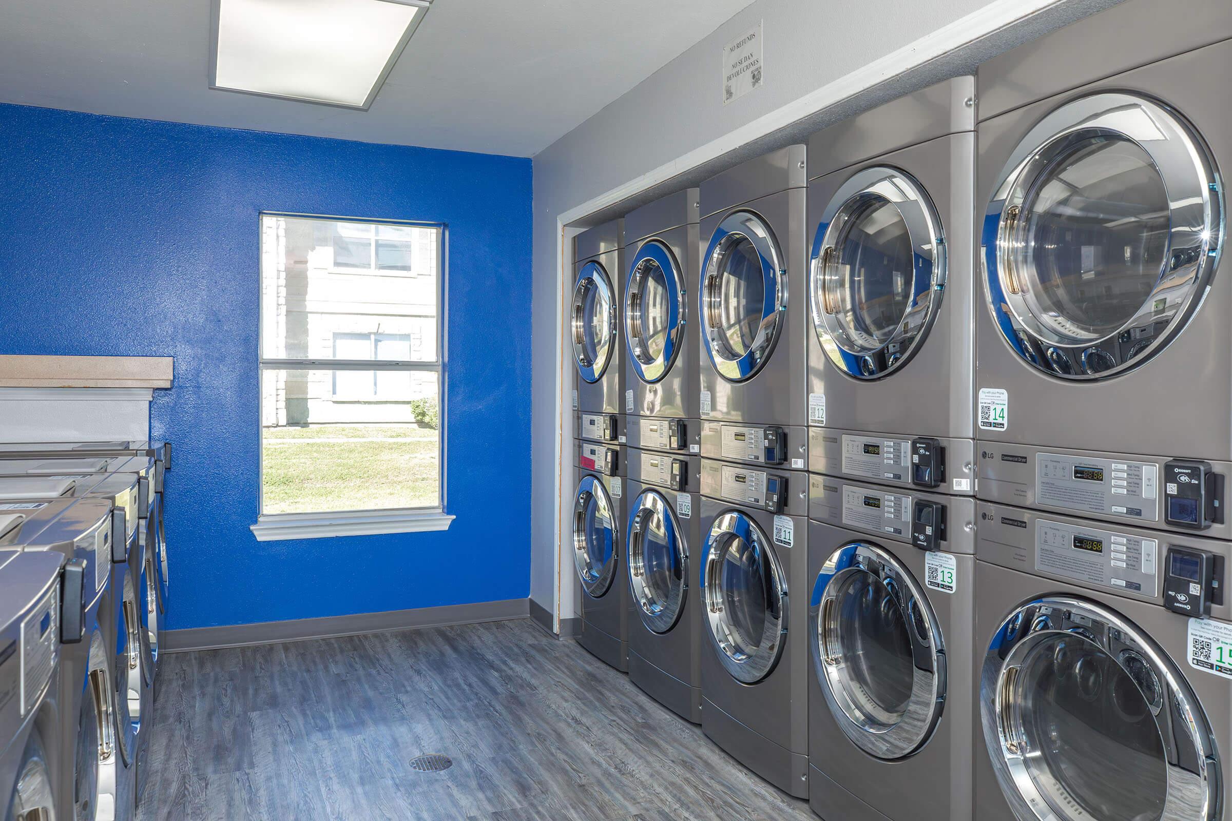 A clean and modern laundry room featuring multiple rows of stainless steel washing machines and dryers. The walls are painted blue, and there is a window allowing natural light to enter. The flooring is a wood-like laminate, and the space is well-lit and organized.