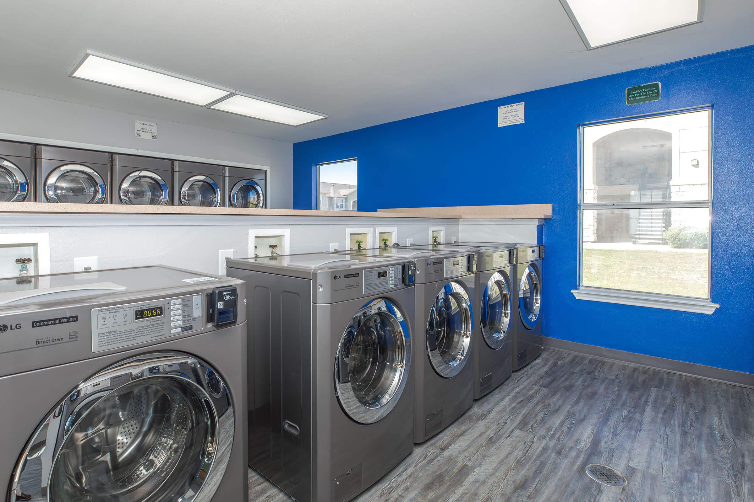 A modern laundry room featuring several silver washing machines and dryers. The walls are painted blue with a light wooden countertop. There are windows allowing natural light, and the space is clean and organized, ideal for laundry chores.