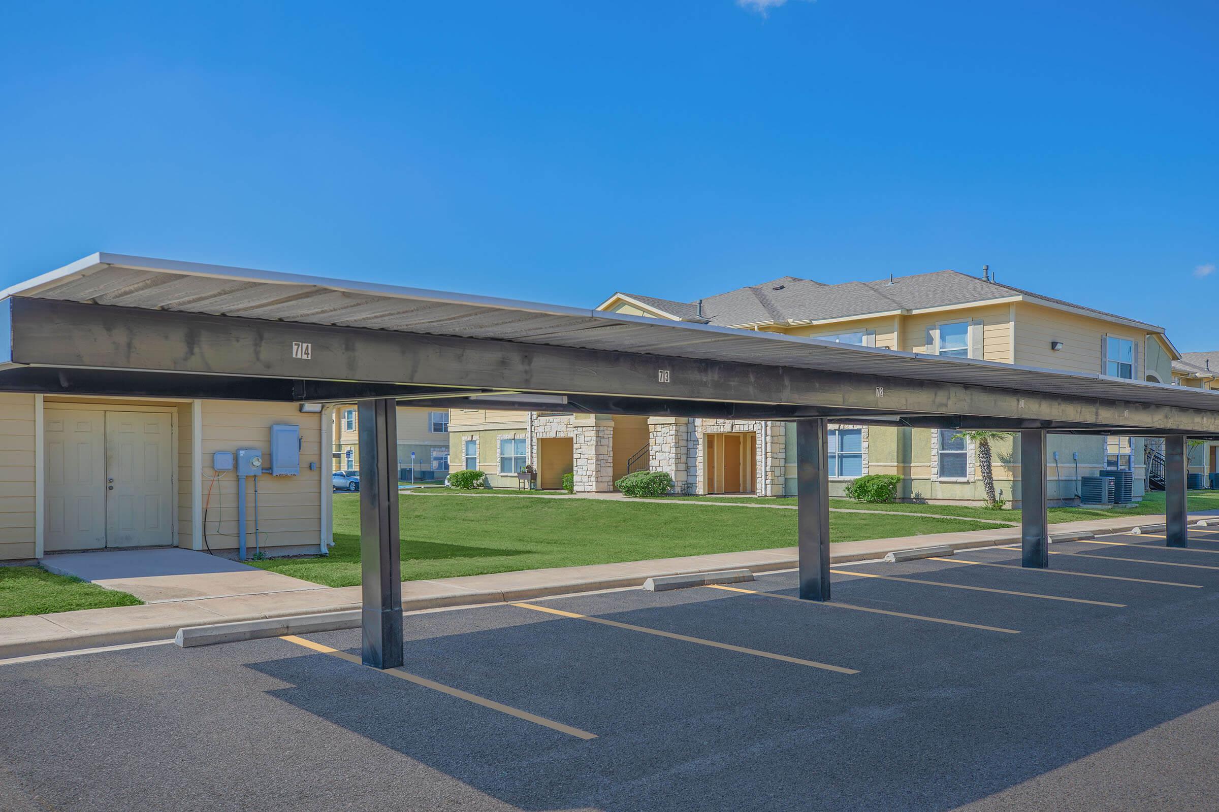 A parking lot with a covered carport structure and multiple parking spaces. In the background, there are apartment buildings with yellow siding, manicured lawns, and clear blue skies. The area is well-maintained and spacious, indicating a residential community.