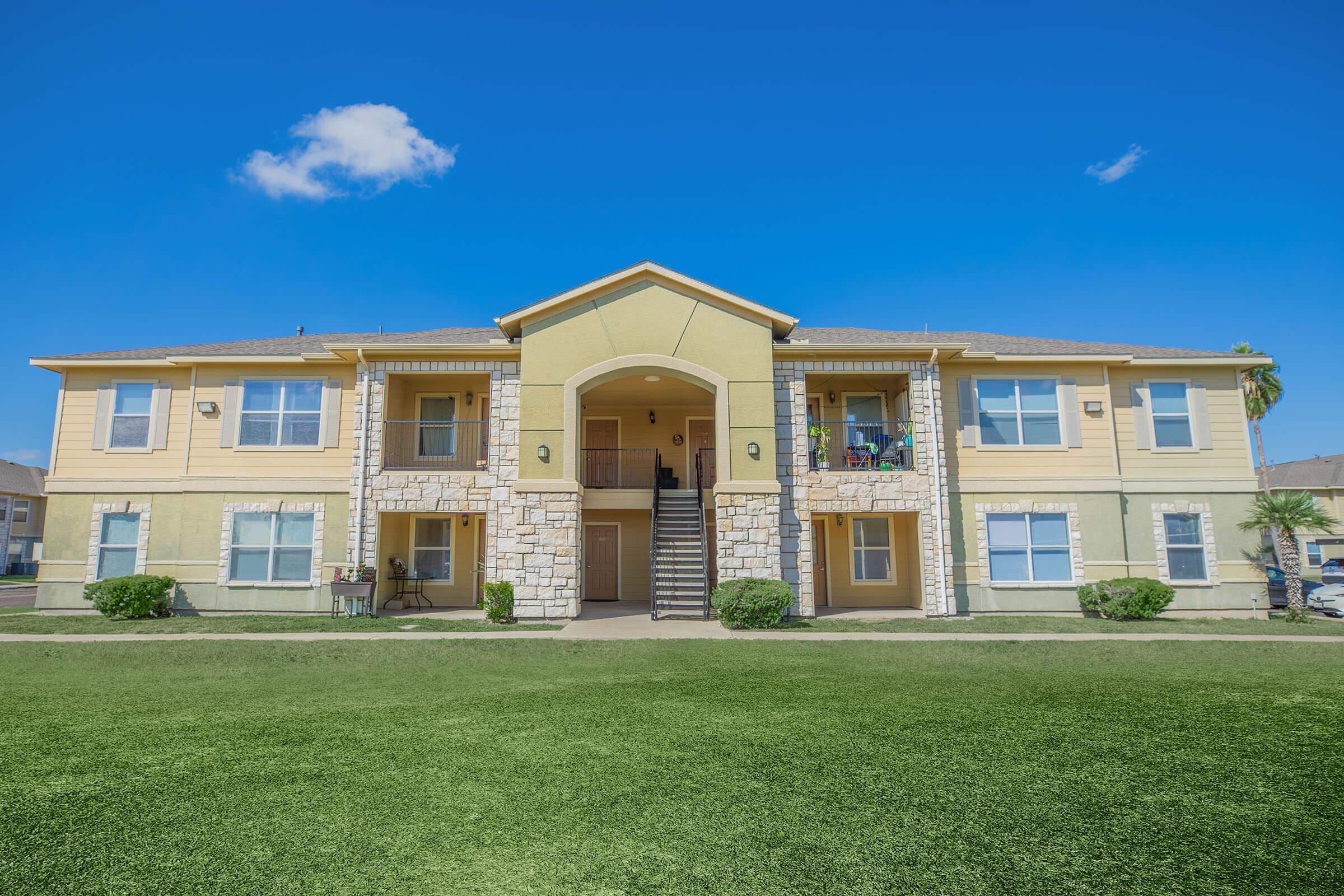 Two-story residential apartment building with a yellow and stone exterior, featuring balconies on the second floor. A well-maintained grassy area is in front, and the sky above is clear and blue with a few clouds.