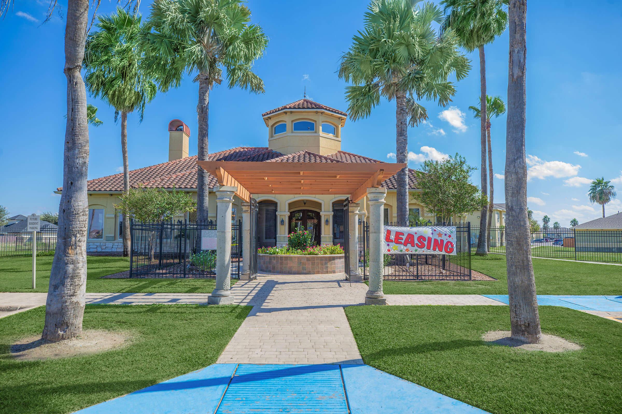 A leasing office building surrounded by palm trees, with a shaded entrance and a colorful "Leasing" banner displayed prominently. Lush green grass and a clear blue sky enhance the warm and inviting atmosphere of the location.