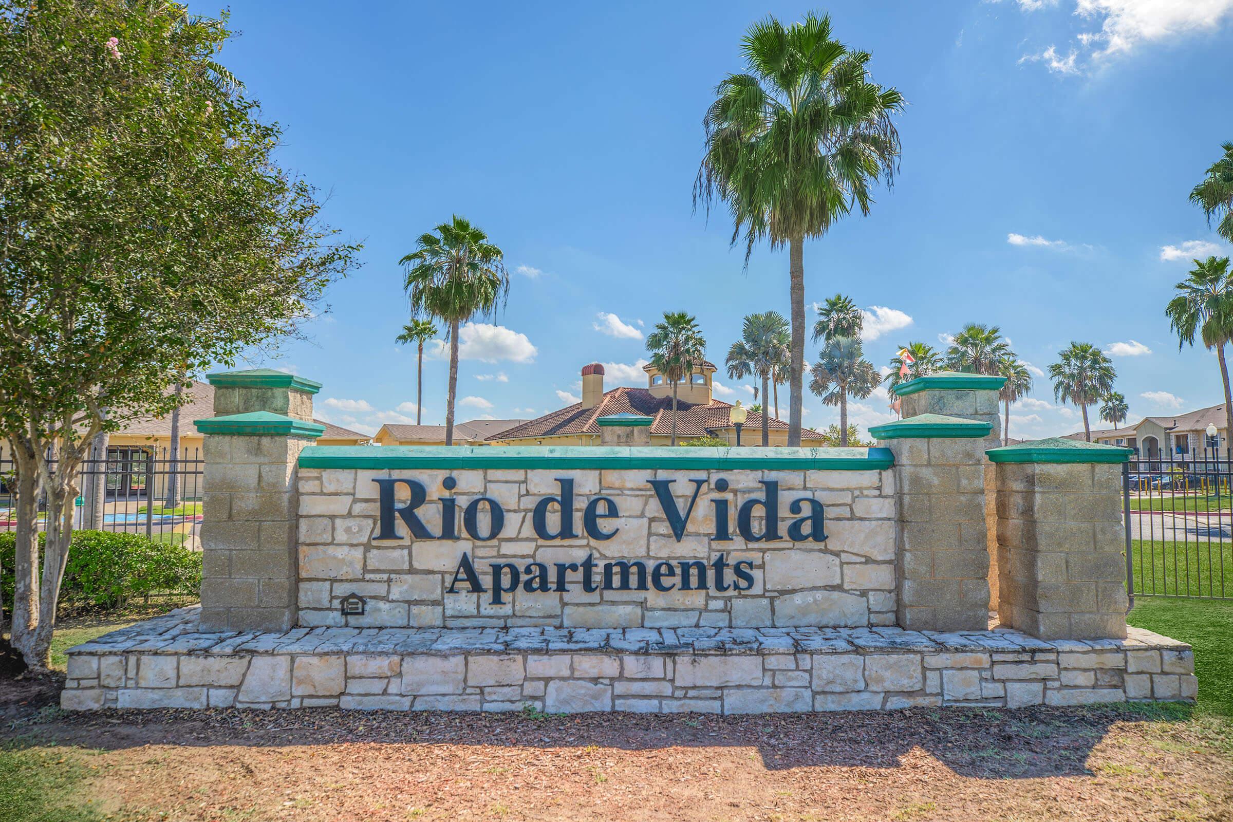 Sign for Rio de Vida Apartments, featuring large black text on a stone background. Surrounding the sign are well-maintained palm trees and a clear blue sky, indicating a sunny day. The apartments are in a residential area with green landscaping.