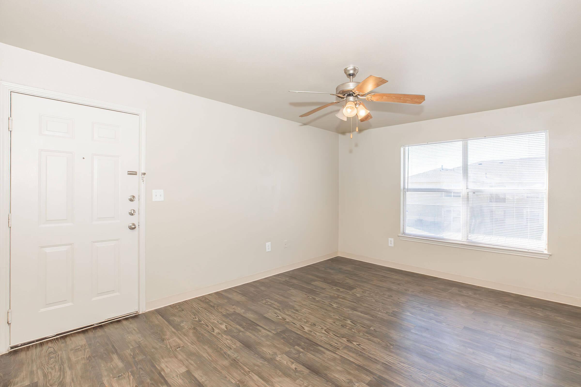 A vacant room featuring a ceiling fan, a large window with natural light, a light-colored wall, and hardwood-style flooring. The entrance door is visible on the left side, and the overall space is clean and ready for furnishings.