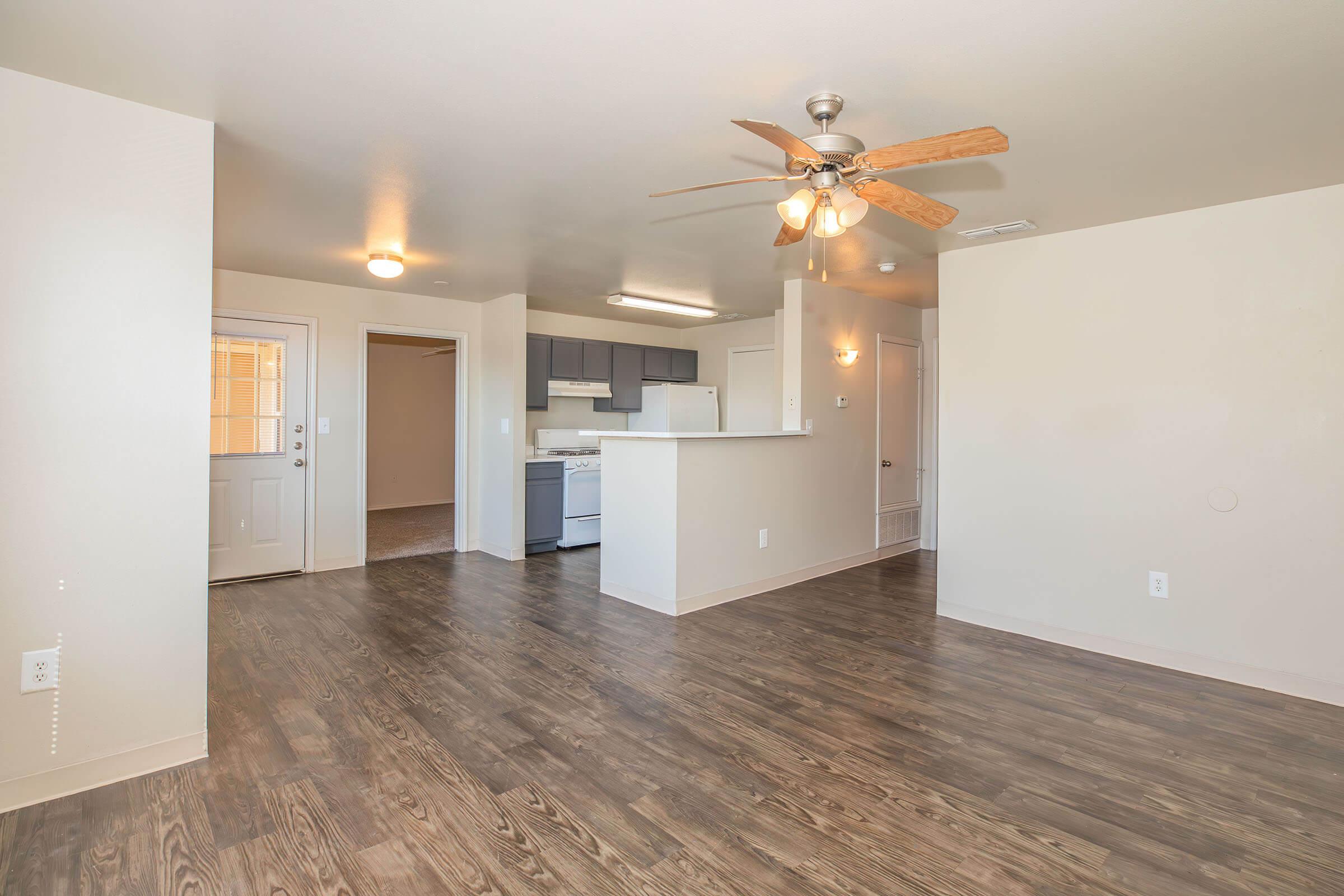 A spacious, well-lit living area in an apartment, featuring a ceiling fan, light-colored walls, and wood-like flooring. The open floor plan connects to a kitchen area with modern appliances, including a stove and refrigerator. A front door leads to the outside, enhancing the inviting atmosphere.